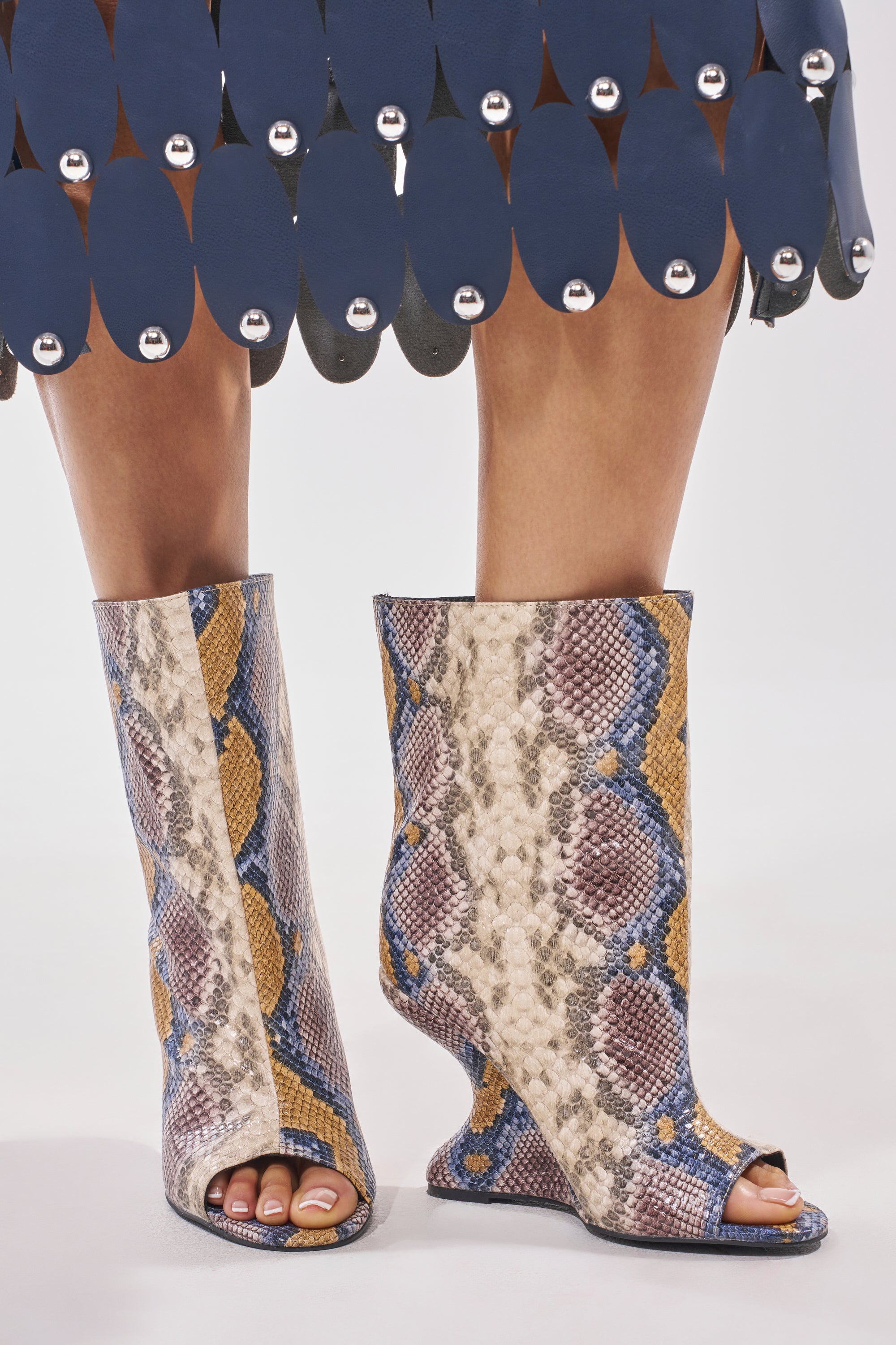 Wearing the AZALEA WANG FAOI SNAKE MULTI OPEN TOE WEDGE BOOTIE—bold blue and yellow snake print booties with striking inverted wedge heels—under a blue skirt with oval cutouts and silver accents. 1