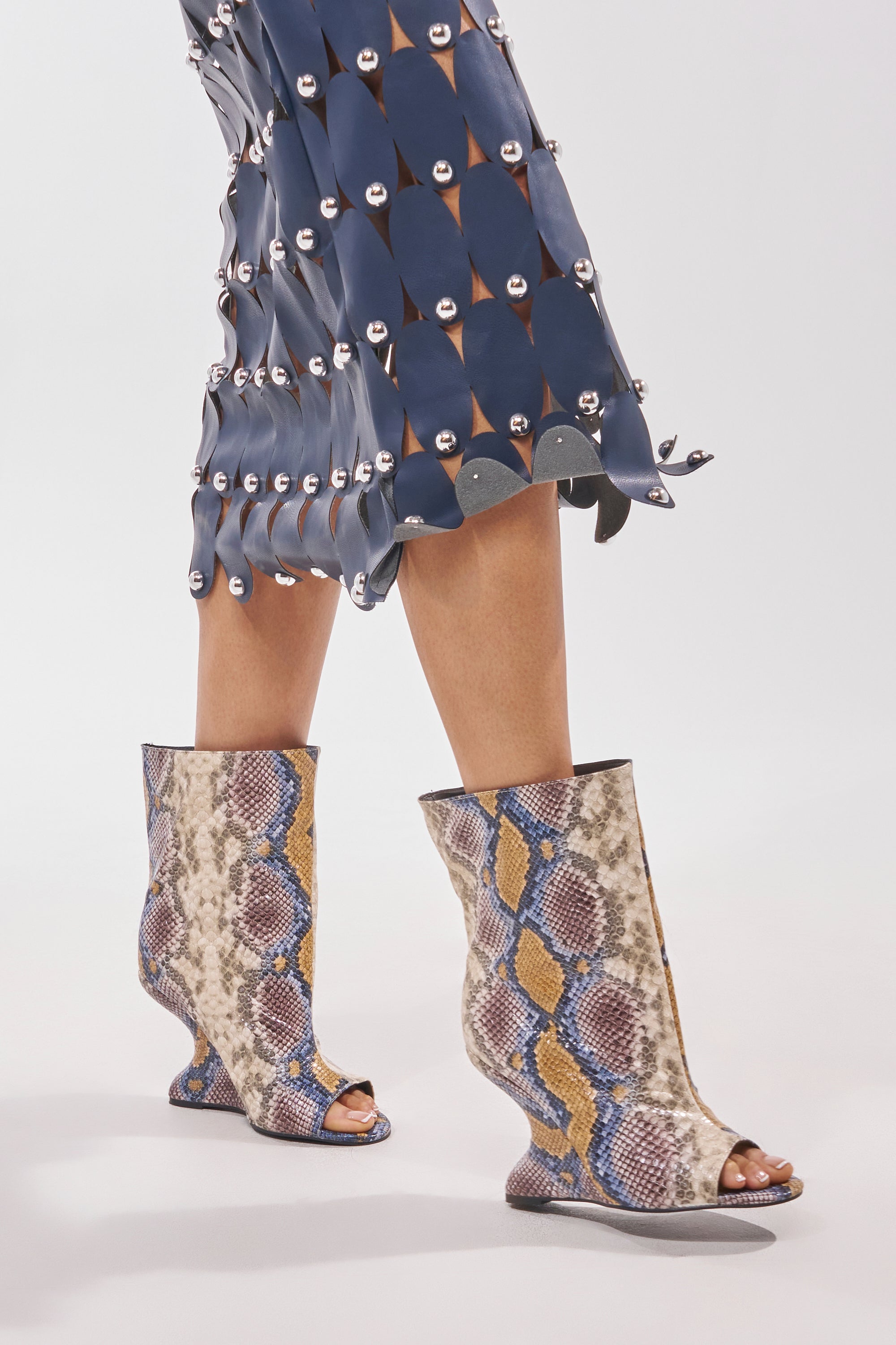 A person wears a blue skirt with round silver embellishments and AZALEA WANG FAOI SNAKE MULTI OPEN TOE WEDGE BOOTIE. The faux leather booties have a snake print and inverted wedge heel. The background is plain and light-colored. 2