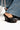 Wearing flared jeans, someone showcases the AZALEA WANG FASCIA BLACK FRINGE WEDGE SANDAL. These vegan leather shoes have a stylish open-toe design, black fringe with silver chain embellishments, and a comfort cushion insole for all-day wear.