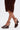 A person wearing a ruched brown faux leather skirt and AZALEA WANG FASCIA CHOCOLATE WEDGE FRINGE SANDALS, featuring open toes, ankle straps, and fringe details, stands with legs crossed against a plain white background.