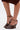 A person wears the AZALEA WANG FASCIA CHOCOLATE WEDGE FRINGE SANDAL, featuring brown faux leather, fringe details, ankle straps, and high wedges. Only the lower legs and feet are shown against a white background.