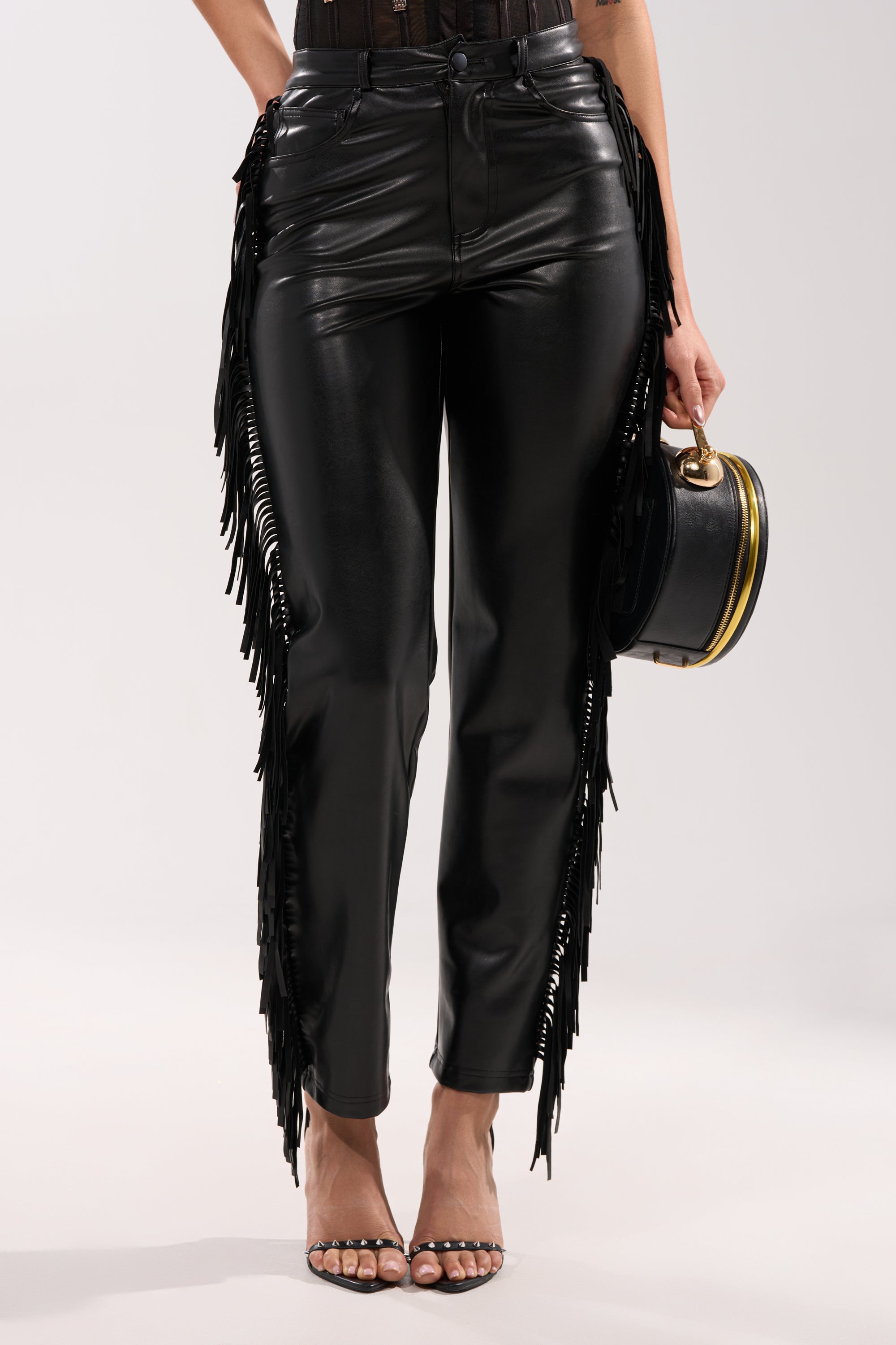 Wearing the LETS GO TO THE RODEO FRINGE FAUX LEATHER PANT in black with side fringe, paired with a sheer black top, strappy heels, and a small round black handbag. 2