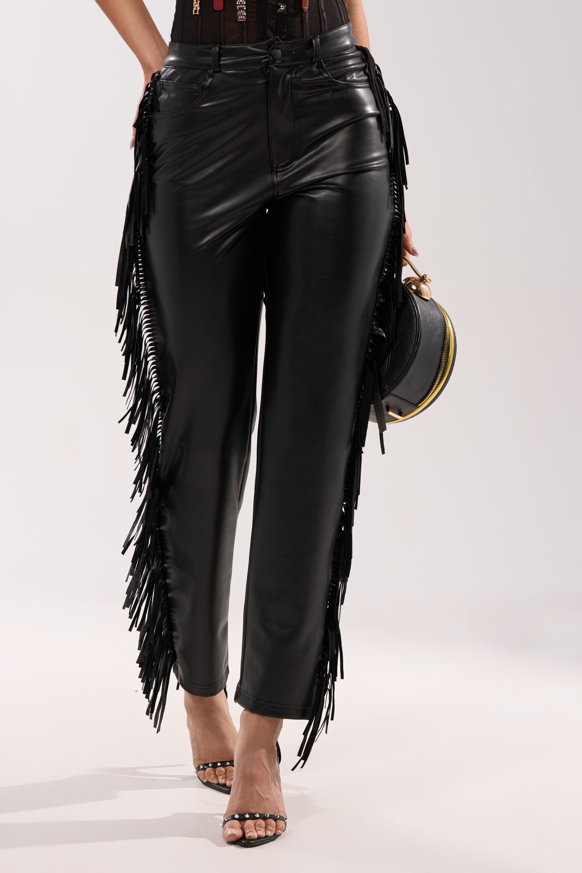 Wearing the LETS GO TO THE RODEO FRINGE FAUX LEATHER PANT with open-toe heels and holding a small black round bag, a person stands against a plain background. 0