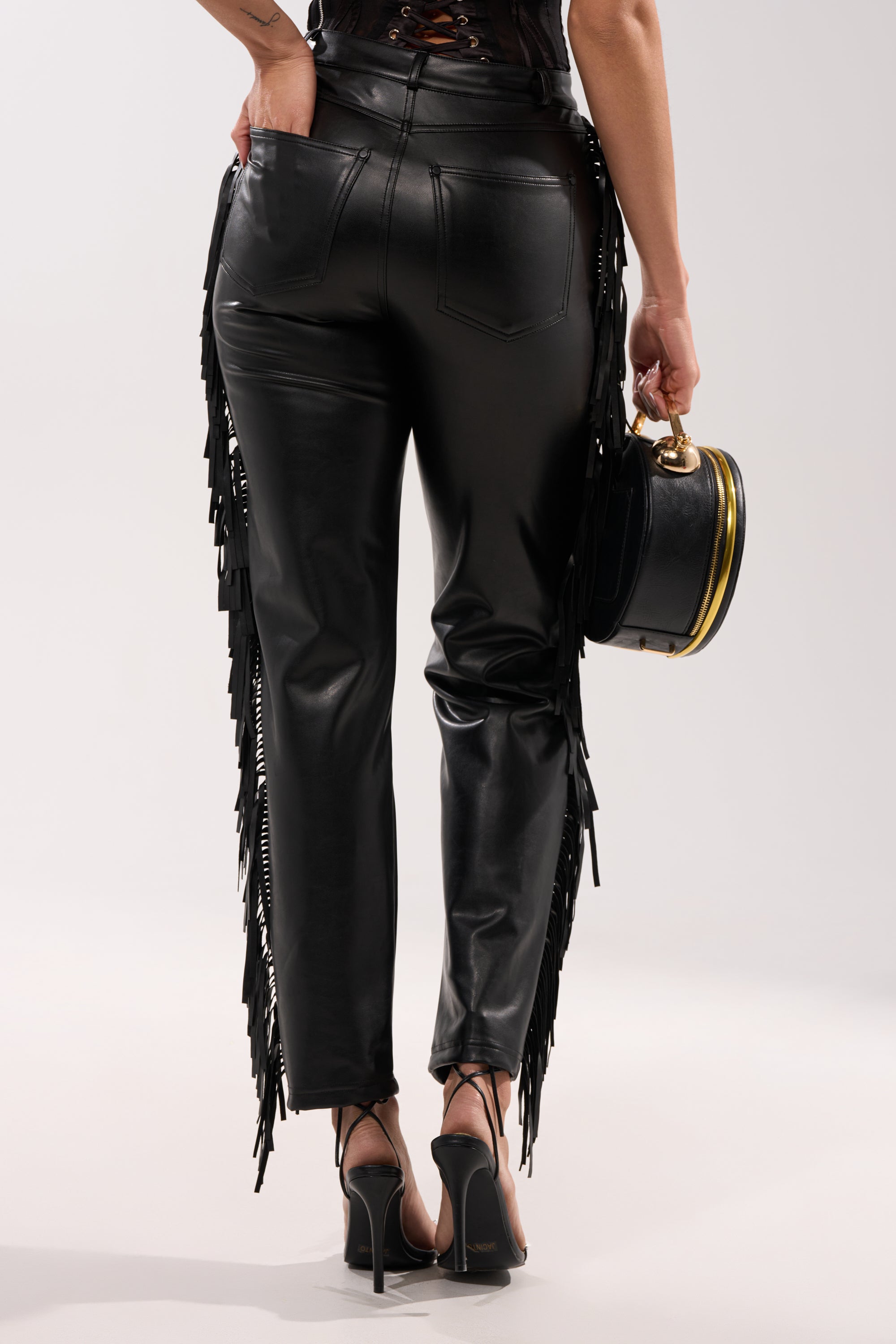 A person is shown from the back wearing LETS GO TO THE RODEO FRINGE FAUX LEATHER PANT with black high-heeled sandals and a small round black handbag, set against a plain light background. 4
