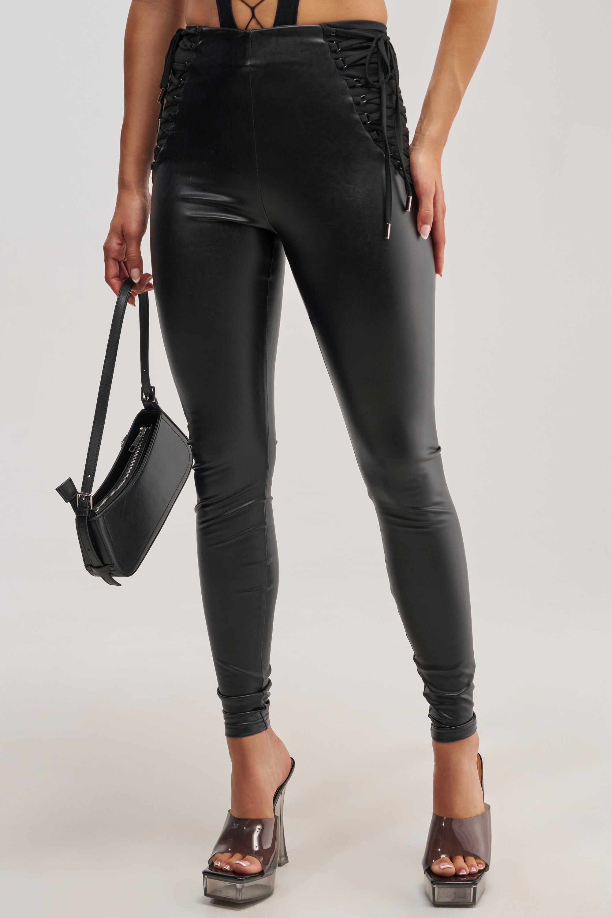 A person wearing the SEXY AS HELL LACE UP LEGGING, styled with a small black handbag and clear high-heeled shoes, stands against a plain background.
