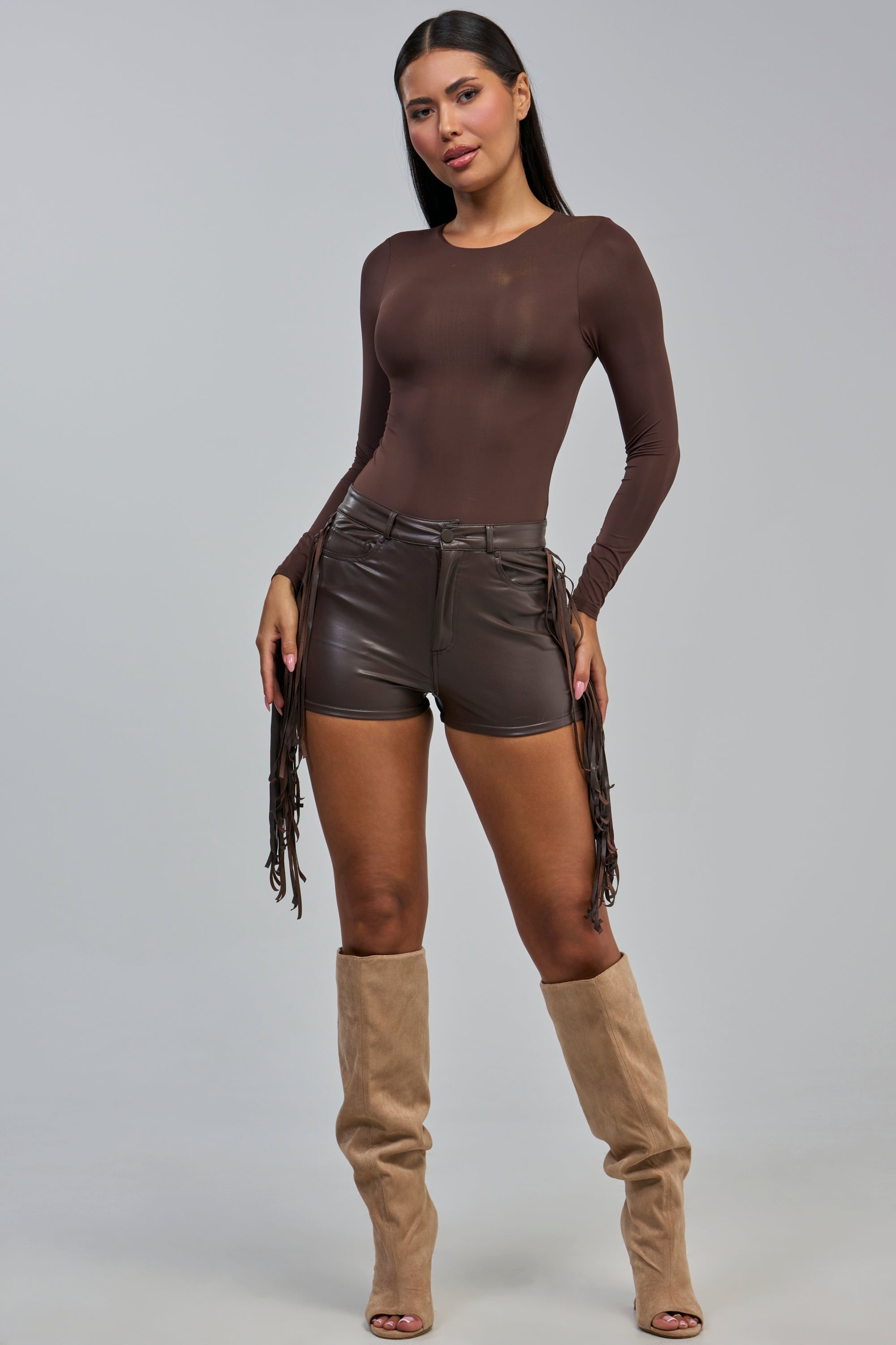 A woman poses against a plain gray background, channeling effortless western style in the RODEO CHIC FAUX LEATHER SHORT, paired with a brown long-sleeve top and beige knee-high peep-toe boots.