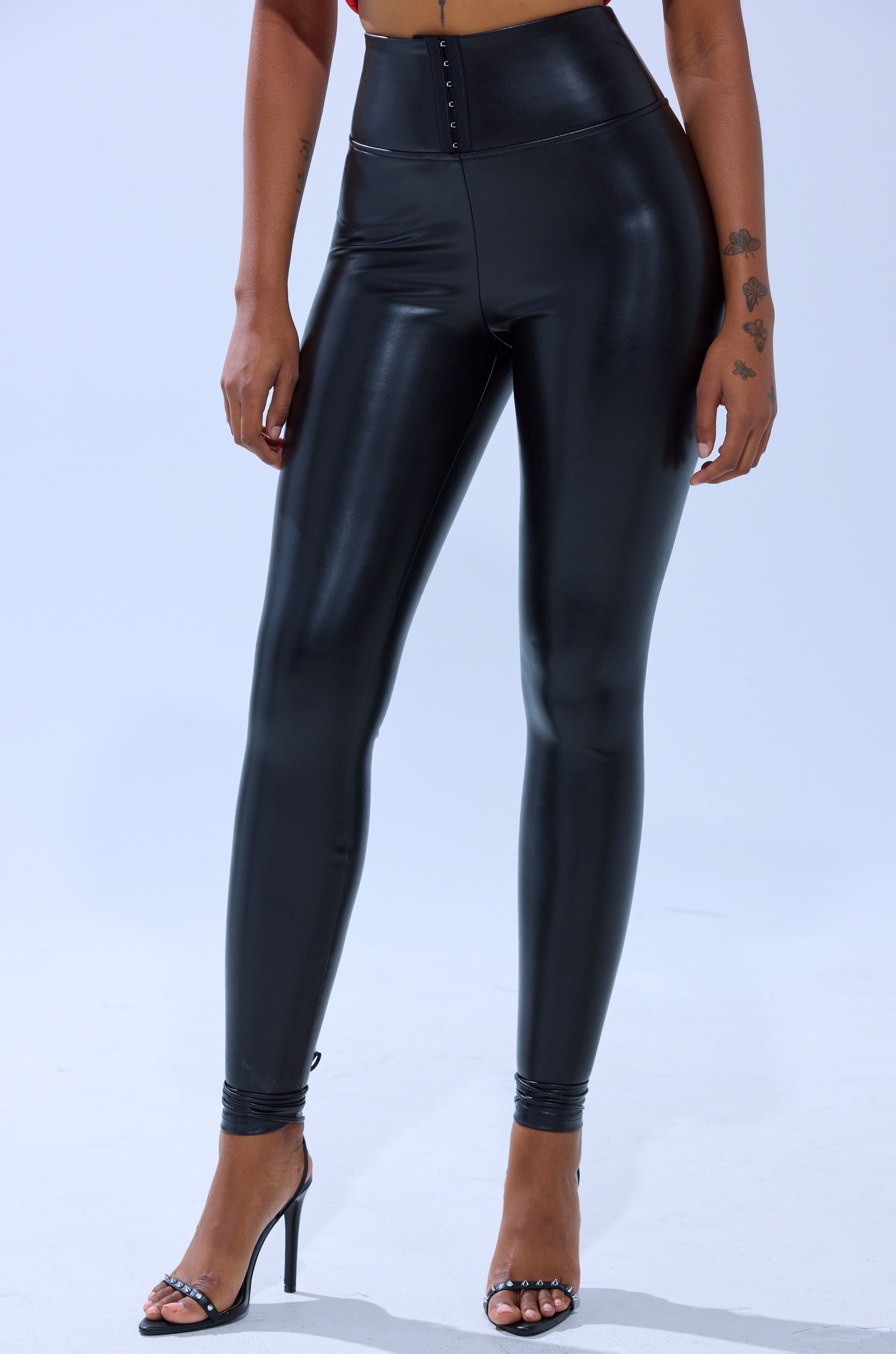 A person stands against a plain light background wearing SLIM THICC CORSET LEGGINGS—high-waisted, black, shiny faux leather—and open-toe heels. Tattoos are visible on their left hand and wrist.