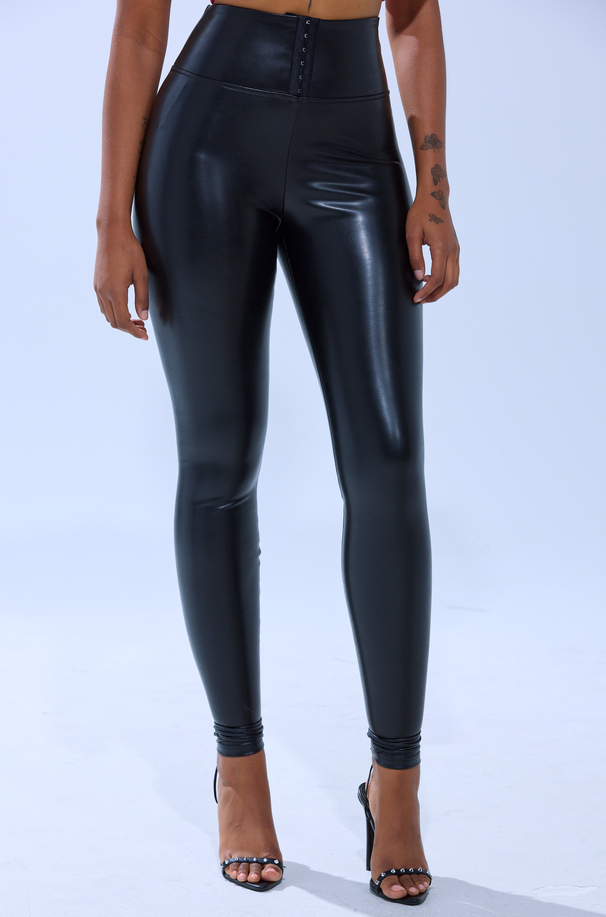 A person wears SLIM THICC CORSET LEGGINGS—high-waisted, shiny black faux leather—paired with black open-toe heels, standing against a plain light background with only the lower body visible.