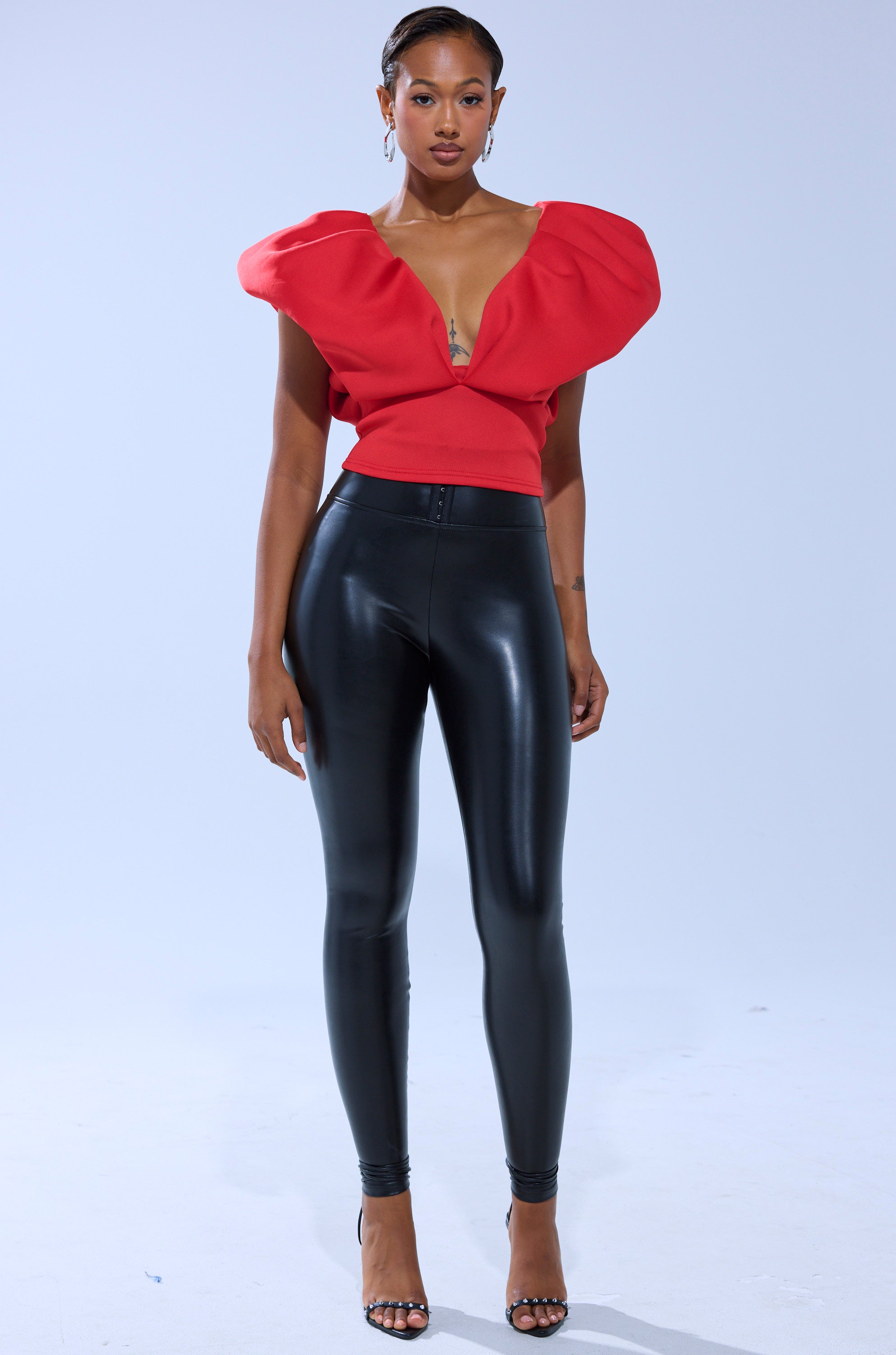 A person stands against a plain background wearing a bold red top with dramatic, sculpted shoulders and a deep V-neckline, paired with SLIM THICC CORSET LEGGINGS in shiny black and black open-toe heels.