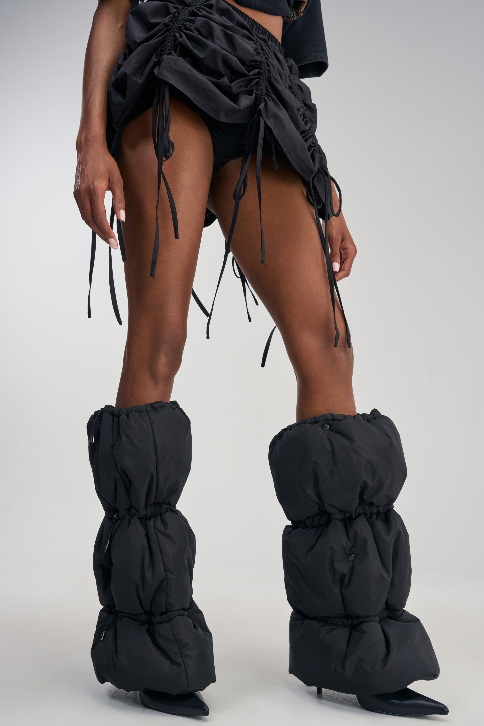 A person is wearing ruched black shorts with dangling ties and oversized, puffy black leg warmers, paired with the AKIRA GOLD FAUSTINE BLACK PUFFER BLACK STILETTO BOOT against a plain background.