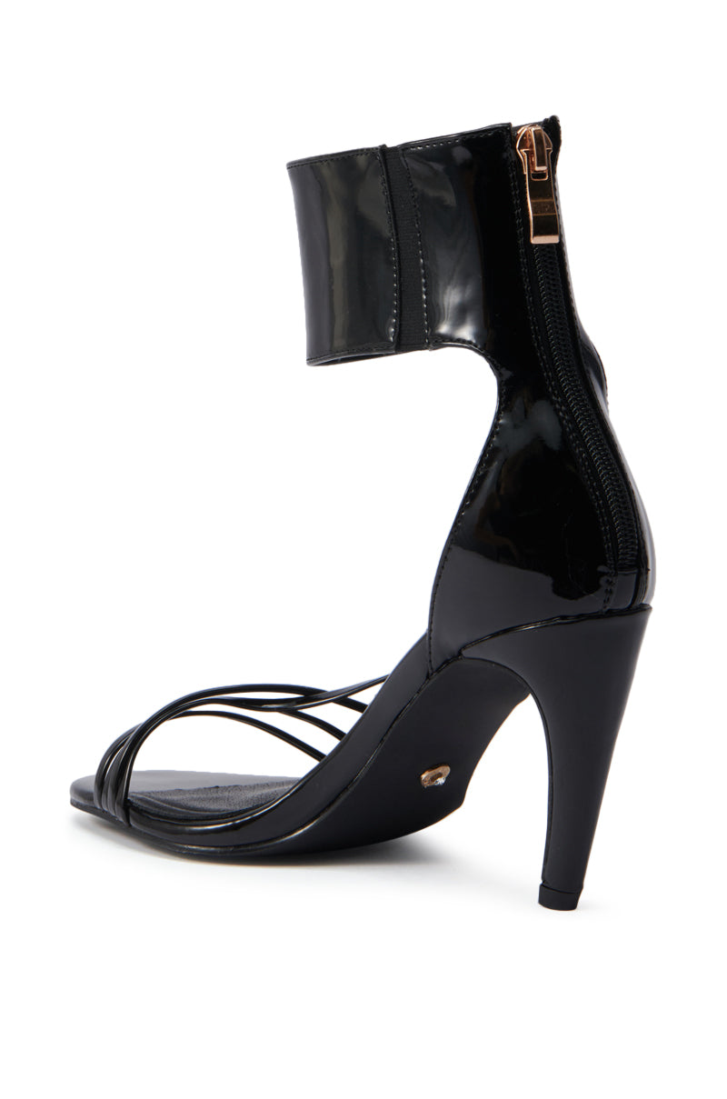 The AZALEA WANG FAWZI Black Banana Heel Cuffed Sandal features thin front straps, an ankle cuff with comfort cushion, a back zipper, and a high heel, shown from a rear side angle on a white background. 4