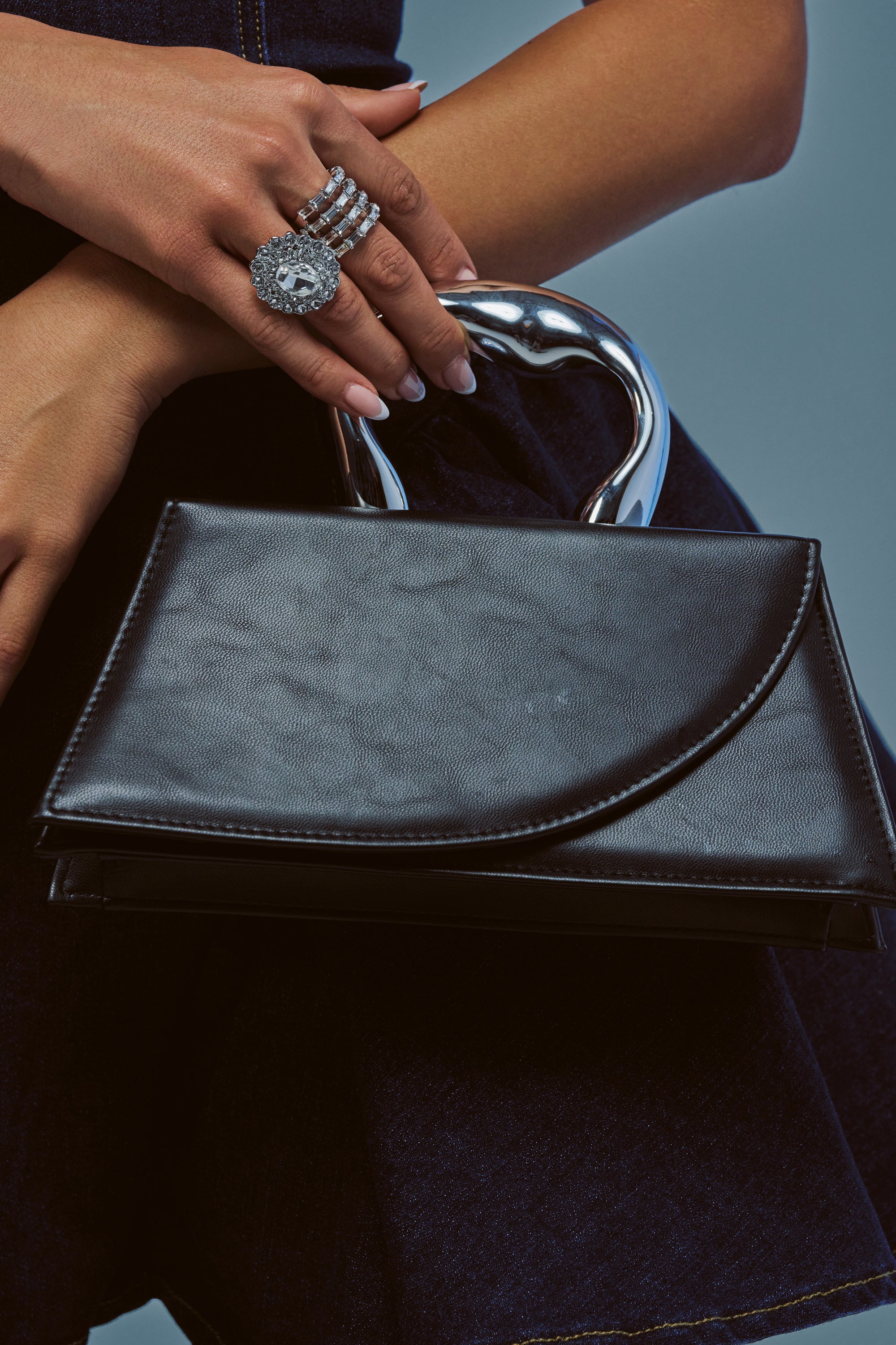 A person holds the YAS QUEEN BAG, a black leather accessory featuring a shiny metallic top handle. They wear a large, decorative ring on their finger against a plain blue background.