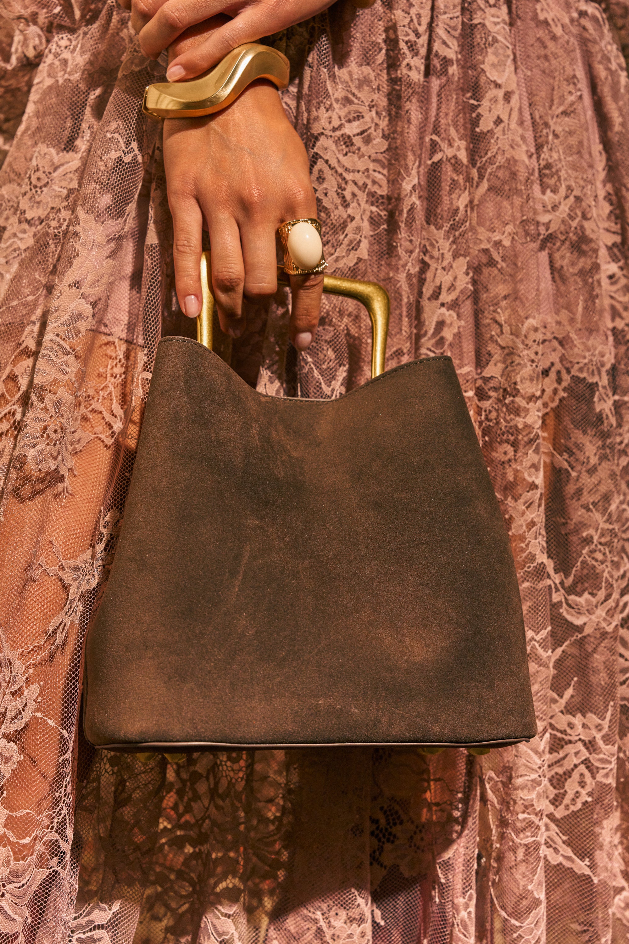 A hand adorned with chunky gold jewelry holds THE BEST BROWN PURSE—a faux suede bucket bag with elegant gold hardware—resting against a pink lace skirt for an effortlessly chic everyday look.