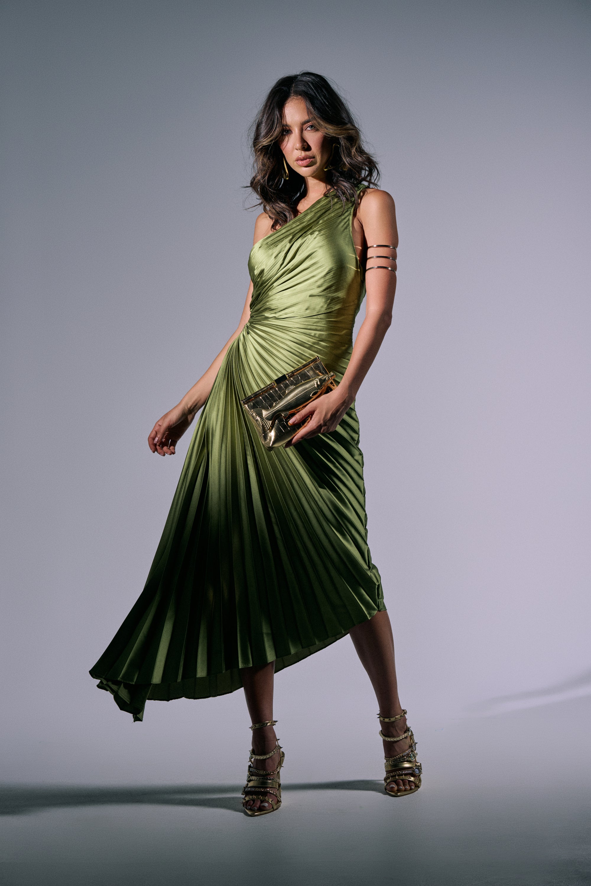 A woman poses confidently in the MIDNIGHT MAVEN SLEEVELESS MAXI DRESS IN GREEN, holding a gold clutch. She wears strappy gold heels and arm cuffs, her wavy hair styled with bold makeup, standing against a plain gray background. 1
