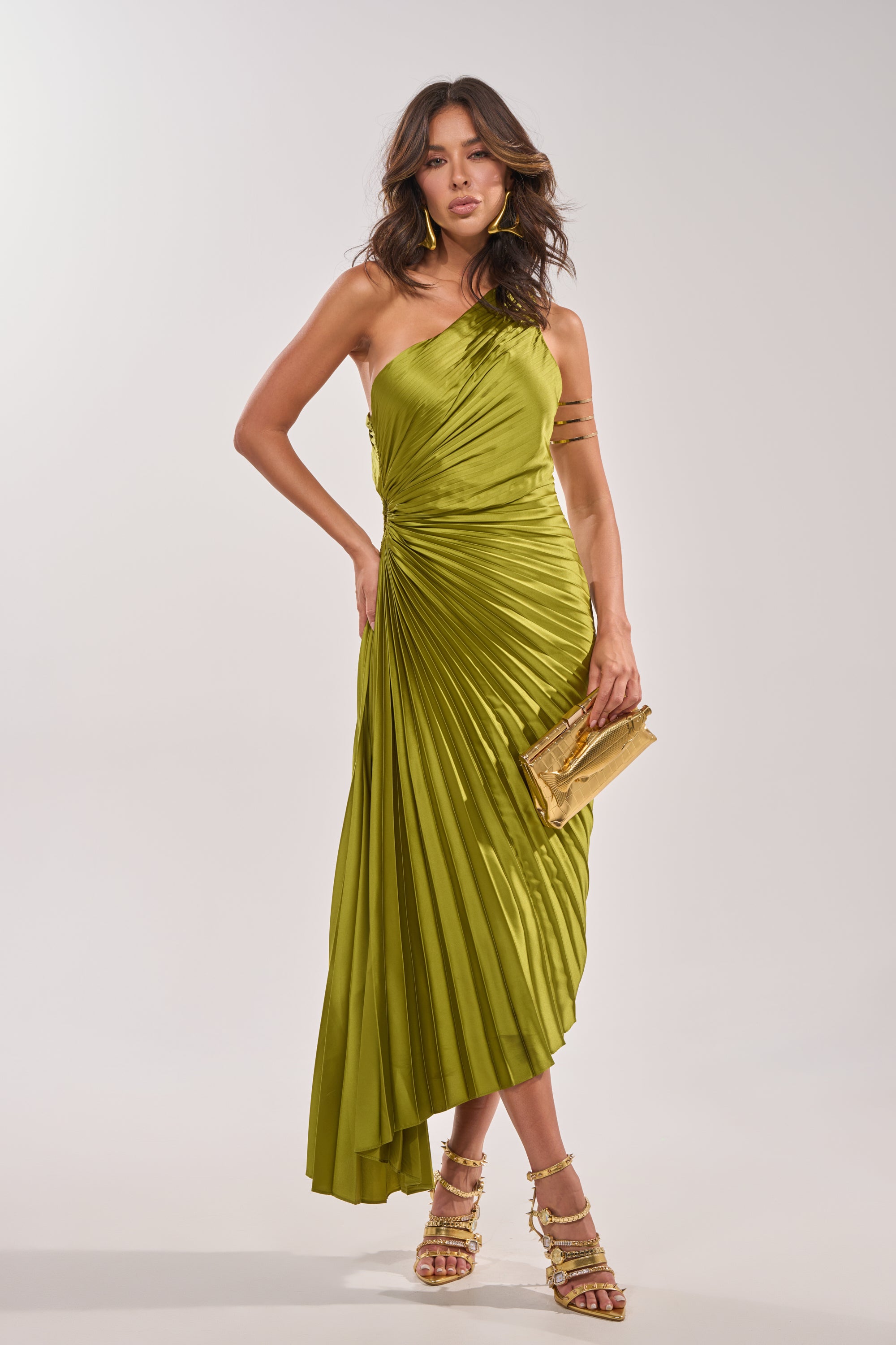A woman wearing the MIDNIGHT MAVEN SLEEVELESS MAXI DRESS IN GREEN, paired with strappy gold heels and a gold clutch, stands confidently against a plain light background.