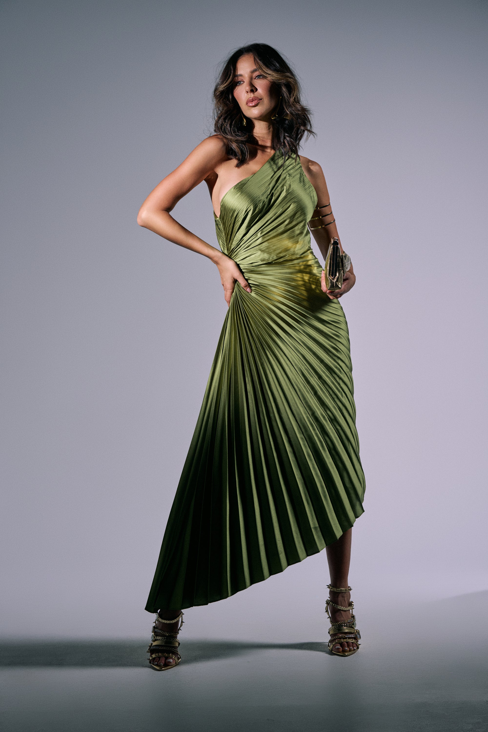 A woman poses confidently in the MIDNIGHT MAVEN SLEEVELESS MAXI DRESS IN GREEN, a shiny pleated satin maxi with an asymmetrical one-shoulder neckline. She holds a clutch and wears strappy heels, her wavy brown hair loose against a neutral background. 2