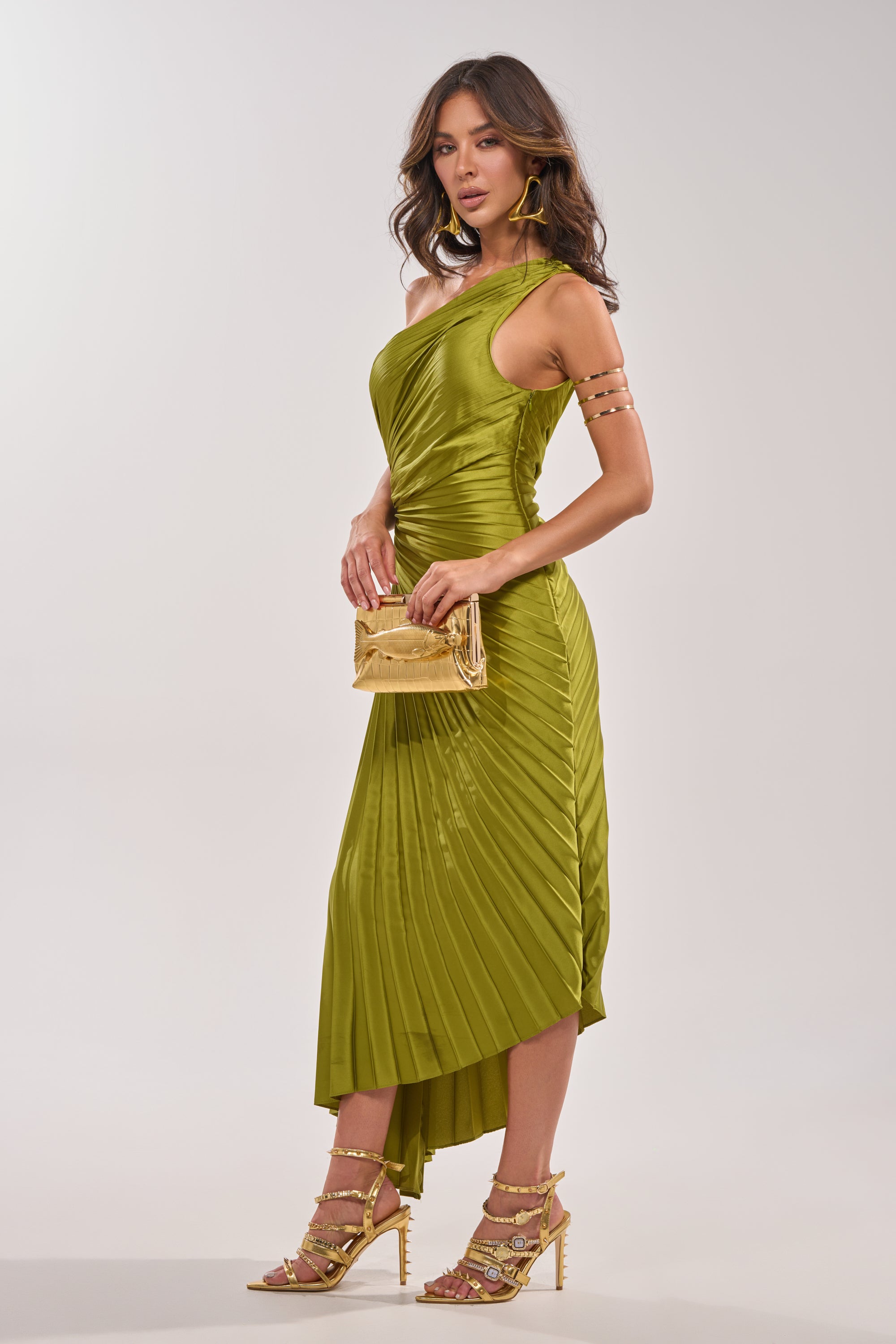 A woman poses against a light background in the MIDNIGHT MAVEN SLEEVELESS MAXI DRESS IN GREEN, featuring a one-shoulder neckline and pleated satin maxi skirt. She completes her look with gold strappy heels, accessories, and a small gold clutch. 5