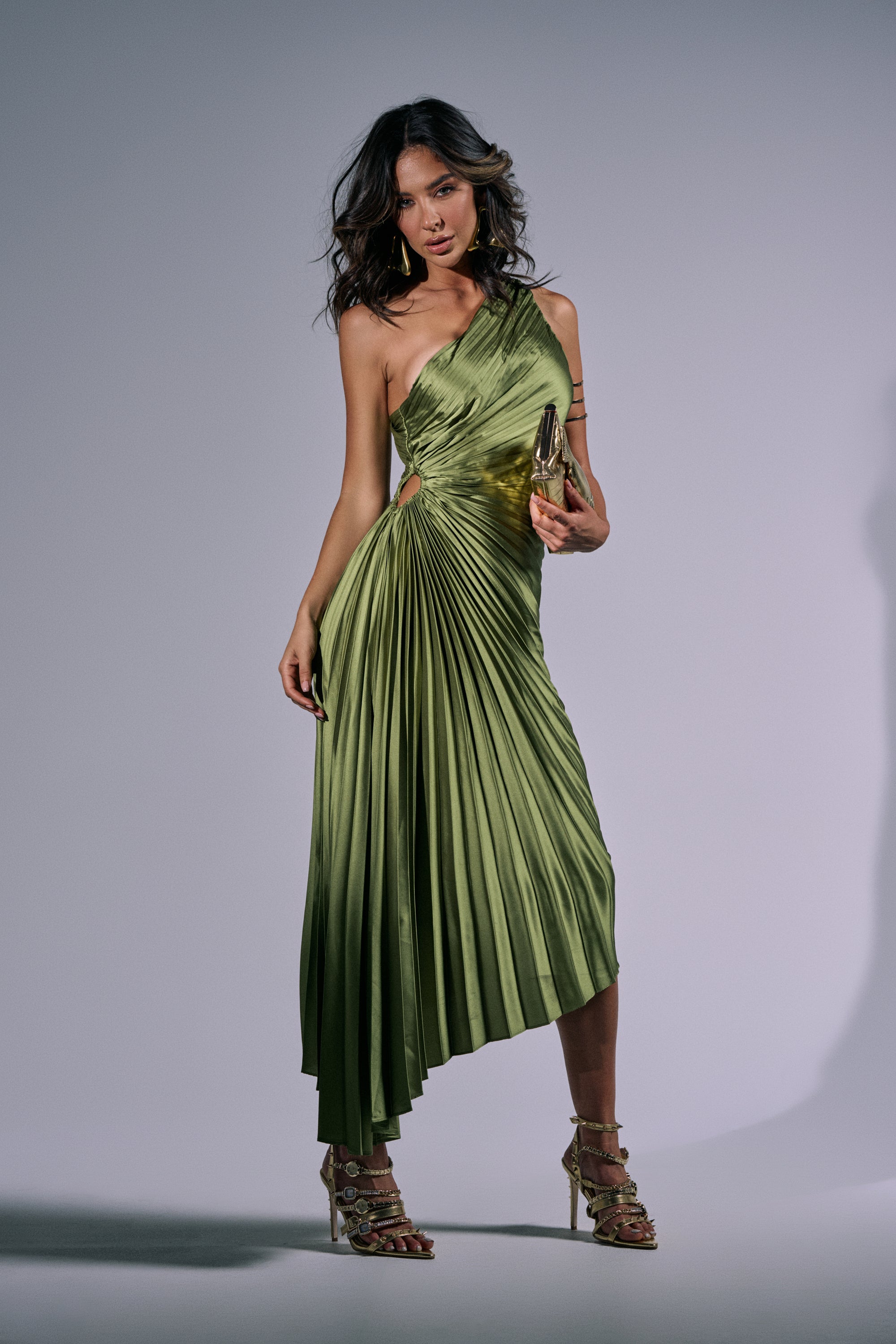 A woman poses in the MIDNIGHT MAVEN Sleeveless Maxi Dress in Green, featuring pleated satin fabric and an asymmetrical one-shoulder neckline. She accessorizes with a gold clutch and strappy heels against a softly lit, plain background. 3