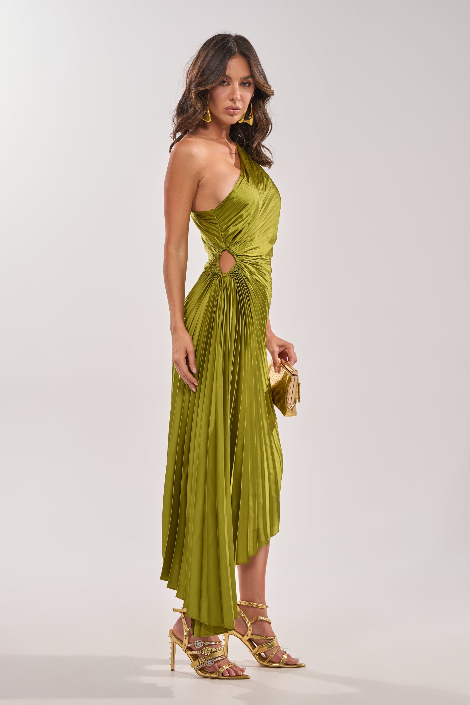 A woman poses in a studio wearing the MIDNIGHT MAVEN SLEEVELESS MAXI DRESS IN GREEN, featuring a shiny finish, one-shoulder neckline, side cutouts, and draped details. She accessorizes with a gold clutch, gold heels, and statement earrings. 6