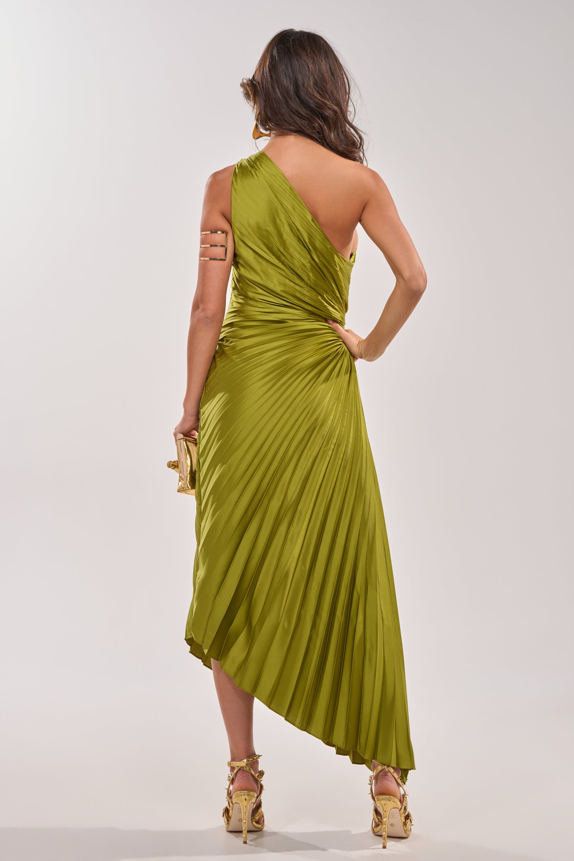 A woman, facing away, wears the MIDNIGHT MAVEN Sleeveless Maxi Dress in green with pleats and a one-shoulder neckline. She holds a gold clutch and sports gold strappy heels against a plain light background. 7