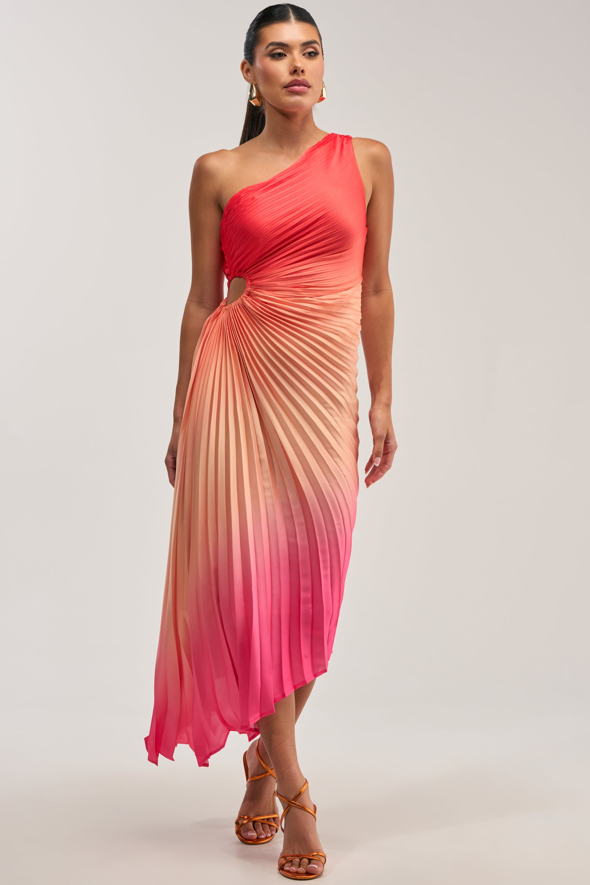 A woman stands against a plain background wearing the WILD CARD One Shoulder Ombre Midi Dress in Pink, featuring delicate pleating and a pink-to-orange gradient, paired with strappy heels.