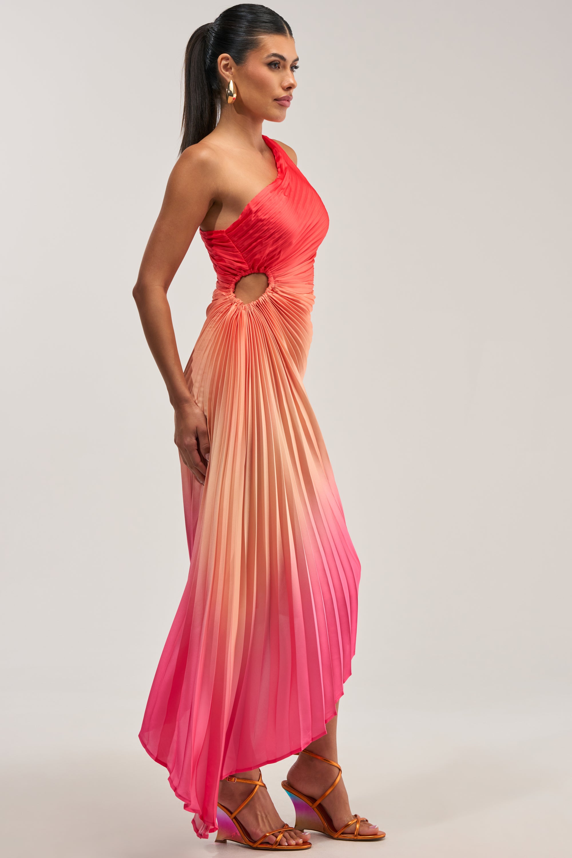 Wearing the WILD CARD ONE SHOULDER OMBRE MIDI DRESS IN PINK—a pleated satin, ombre pink and orange one-shoulder dress with a side cutout—she pairs it with orange strappy heels and gold earrings, standing against a plain background.