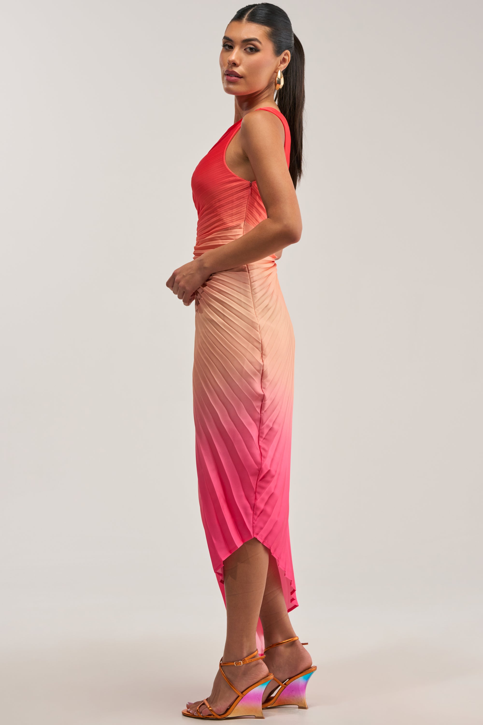 A woman wearing the WILD CARD ONE SHOULDER OMBRE MIDI DRESS IN PINK, which transitions from red at the top to pink at the bottom, stands sideways with her hair in a ponytail and orange heeled sandals.