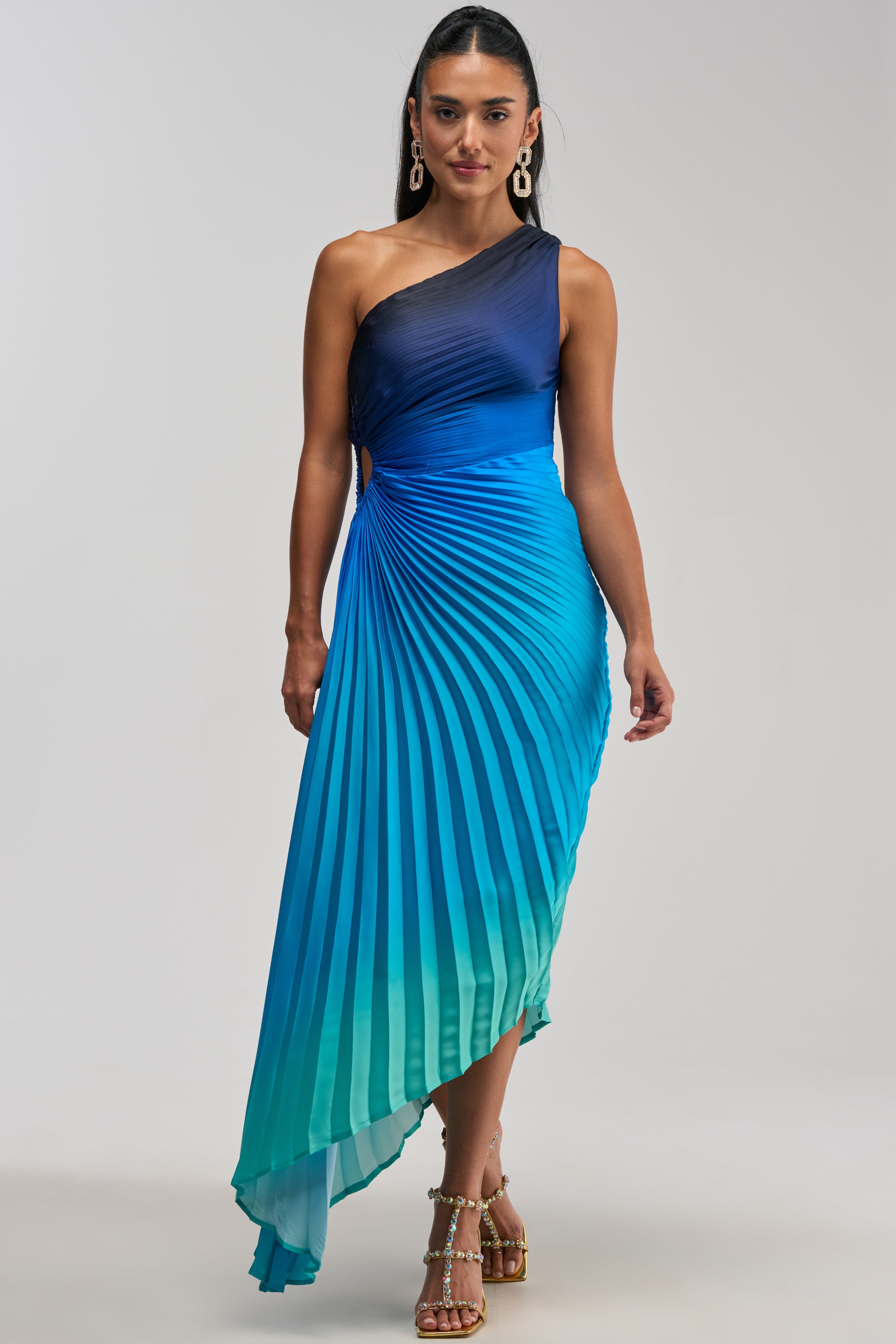 A woman stands against a plain light background wearing the WILD CARD ONE SHOULDER OMBRE MIDI DRESS IN BLUE—a pleated, asymmetrical evening dress featuring a striking blue and teal gradient—styled with dangling earrings and strappy heels.