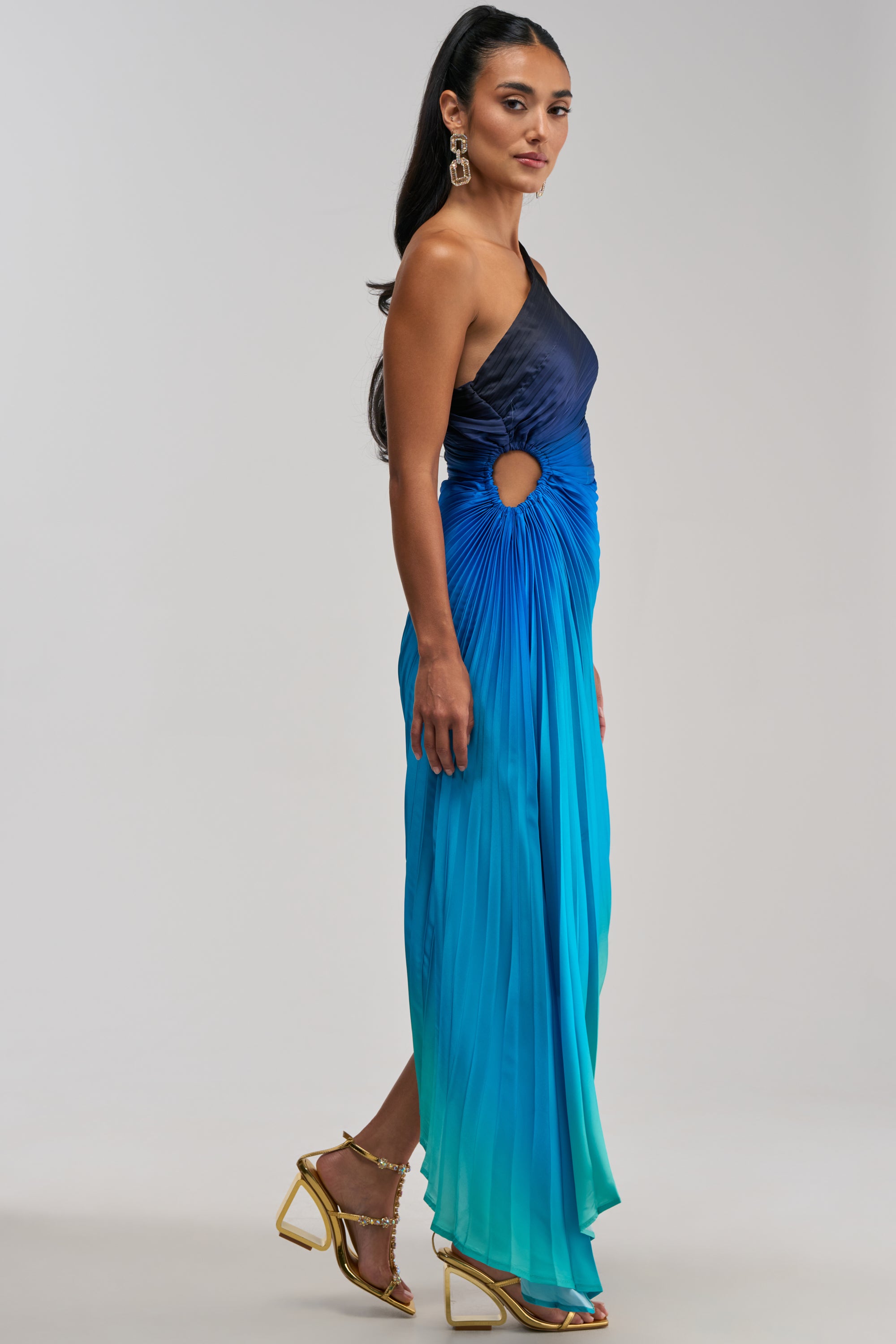 A woman wearing the WILD CARD ONE SHOULDER OMBRE MIDI DRESS IN BLUE—a sleeveless, one-shoulder satin midi with pleats and a side cutout—paired with gold earrings and strappy gold heels, stands against a plain light background.