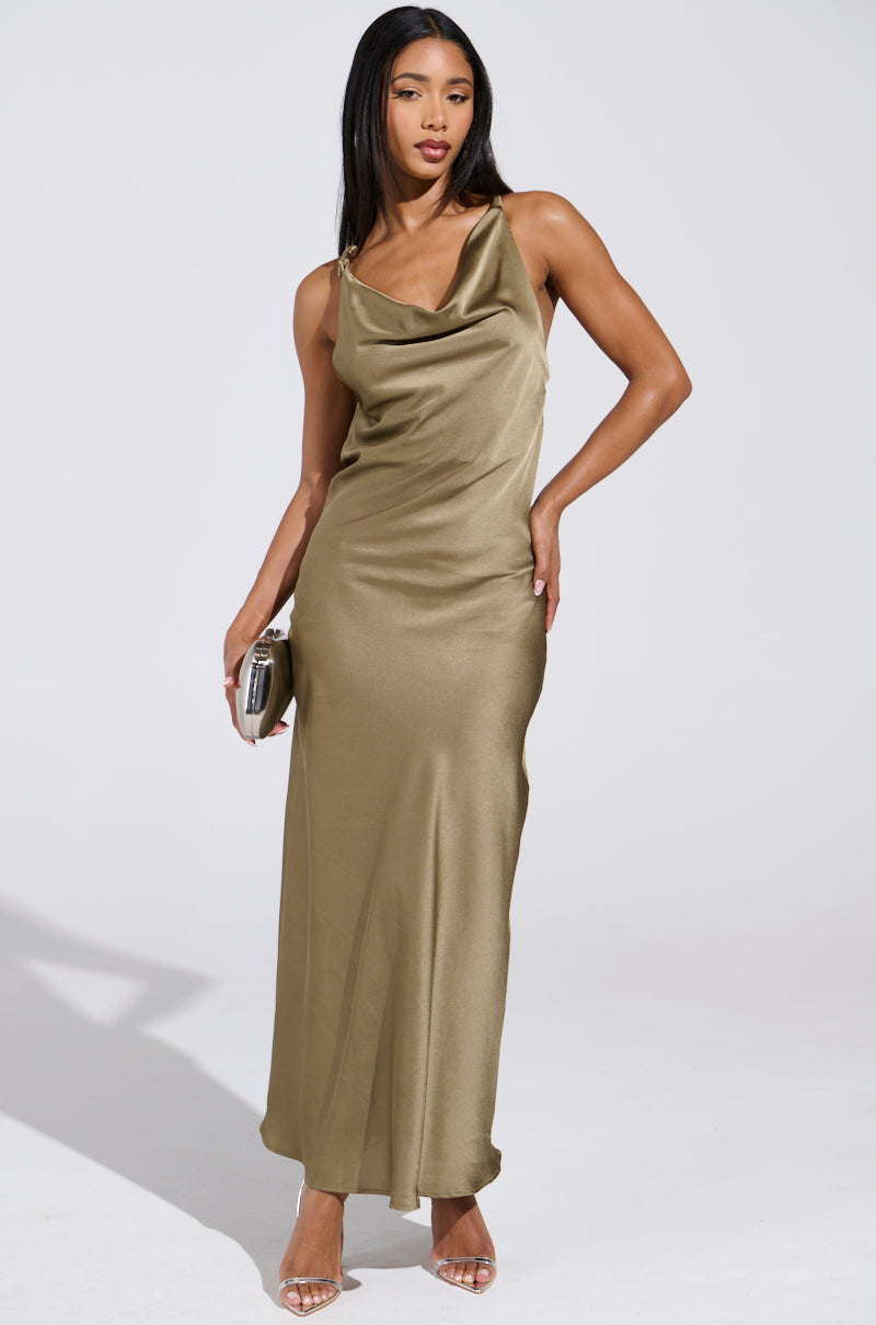 A woman models the BELLA SATIN MAXI DRESS IN OLIVE, featuring a draped neckline and thin straps, ideal for weddings. She accessorizes with a silver clutch and clear high-heeled sandals against a white wall.