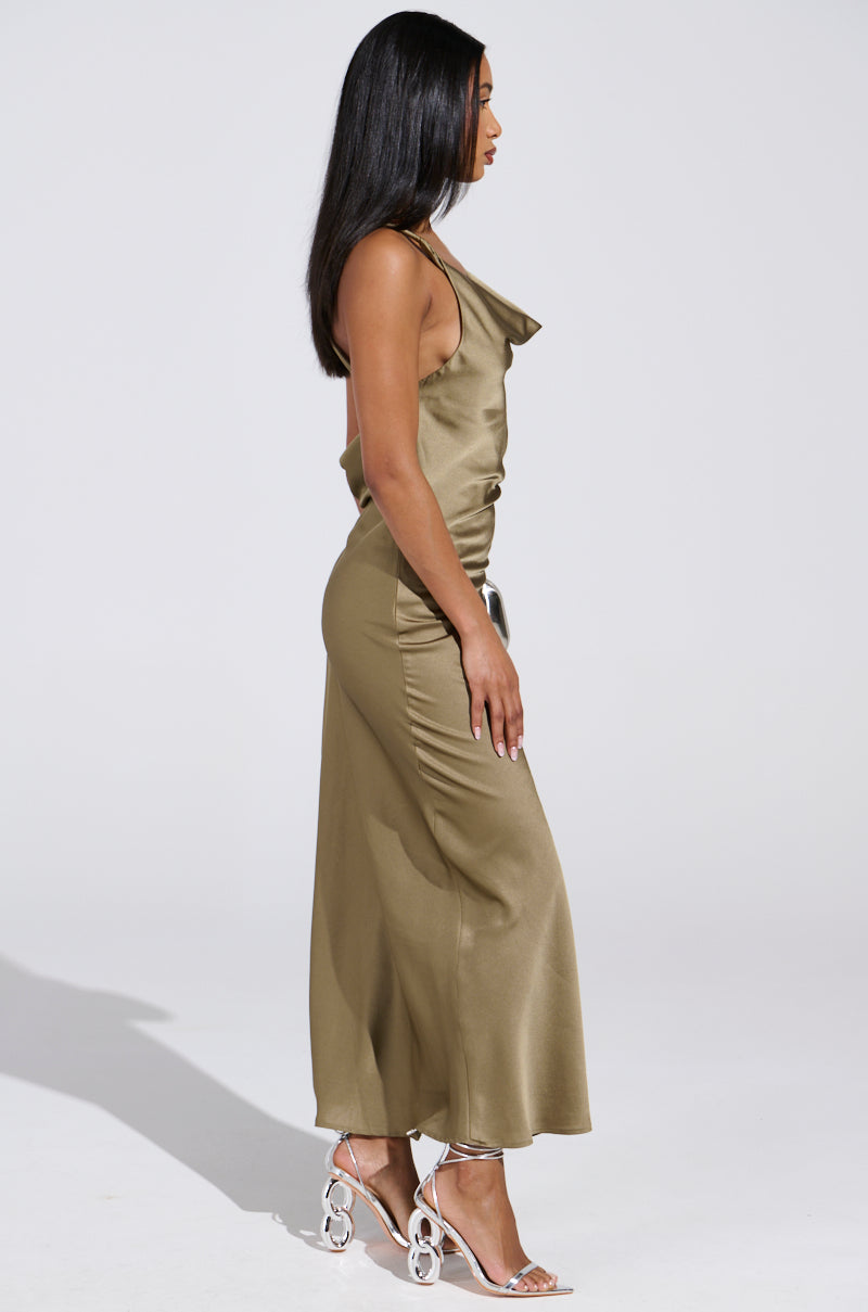 A woman stands against a light background wearing the BELLA SATIN MAXI DRESS IN OLIVE, a sleeveless satin maxi dress, with white strappy high heels. Her long dark hair cascades down as she rests her right hand on her hip—ideal for wedding season.
