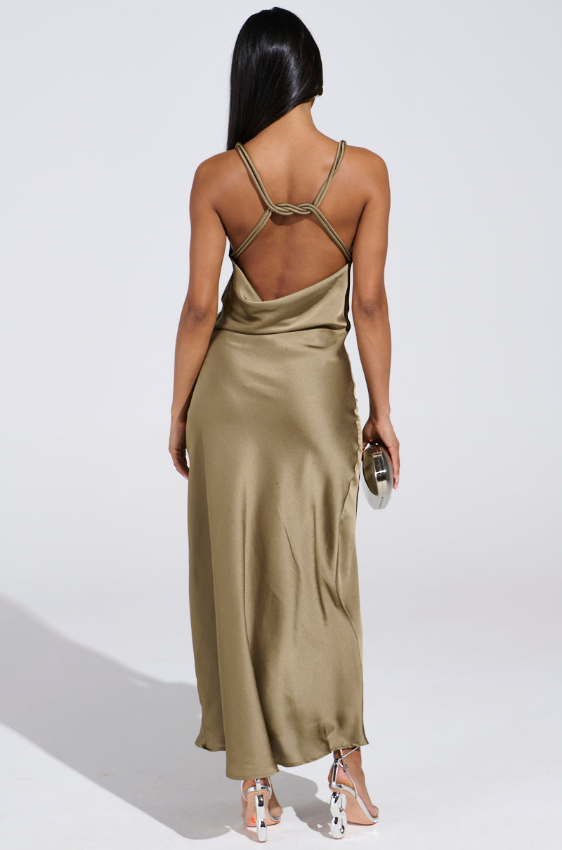 A person with long black hair wears the elegant BELLA SATIN MAXI DRESS IN OLIVE, ideal for wedding season. Holding a small clutch, they stand with their back to the camera, showcasing the dress's draped design and thin straps. Strappy high heels complete this sophisticated look.