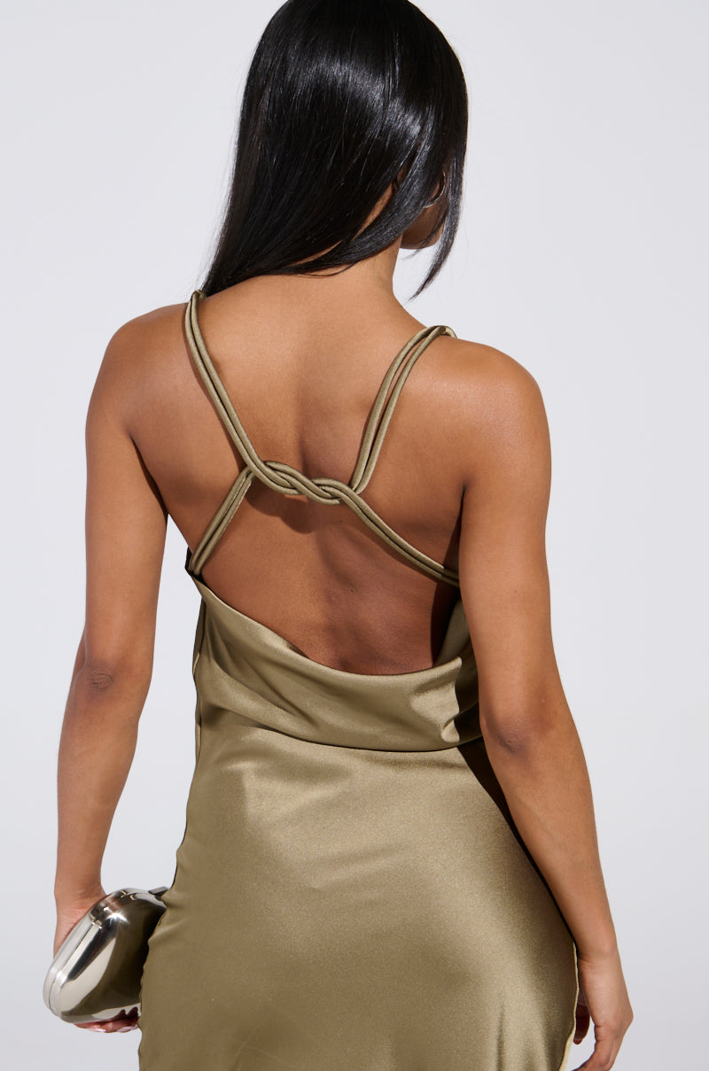 A woman with long black hair, shown from the back, exudes elegance in the BELLA SATIN MAXI DRESS IN OLIVE with crisscross straps—ideal for wedding season. She holds a metallic clutch against a plain background.