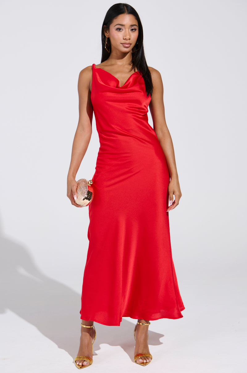 A woman exudes confidence in the BELLA SATIN MAXI DRESS IN RED, featuring a stunning open-back design and cowl neckline. She pairs it with a small, round purse and gold heels. Her straight, loose hair contrasts beautifully against the plain white background.