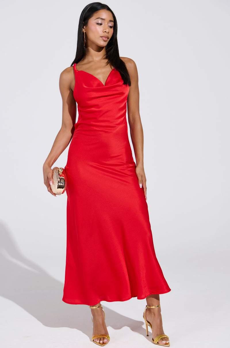 A woman wearing the BELLA SATIN MAXI DRESS IN RED, featuring a sleeveless design and cowl neckline, stands elegantly. She pairs it with a small gold clutch and gold high heels. With straight long hair, she gazes slightly downwards against a plain background, exuding elegance and poise.