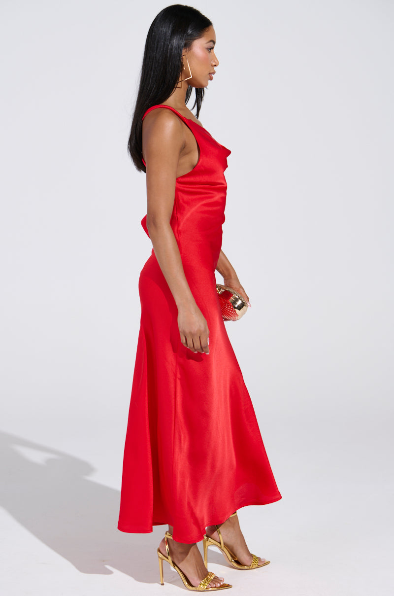 A woman in the BELLA SATIN MAXI DRESS IN RED stands sideways against a plain backdrop. She wears gold high-heeled sandals, holds a small clutch, and her long dark hair falls straight down her back. Hoop earrings adorn her look.