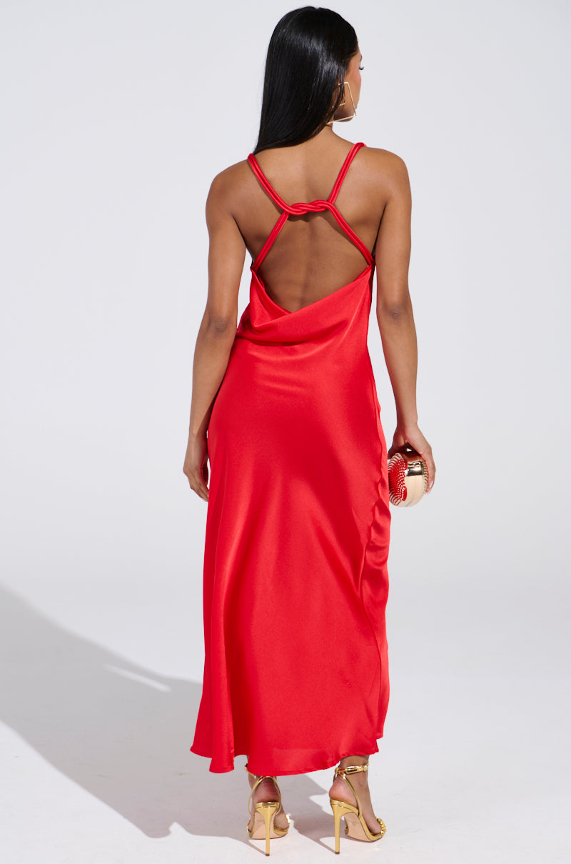 A woman faces away, donning the BELLA SATIN MAXI DRESS IN RED, which showcases an open-back with thin straps. She holds a small gold clutch and wears gold high-heeled sandals. Her long hair is straight and loose.