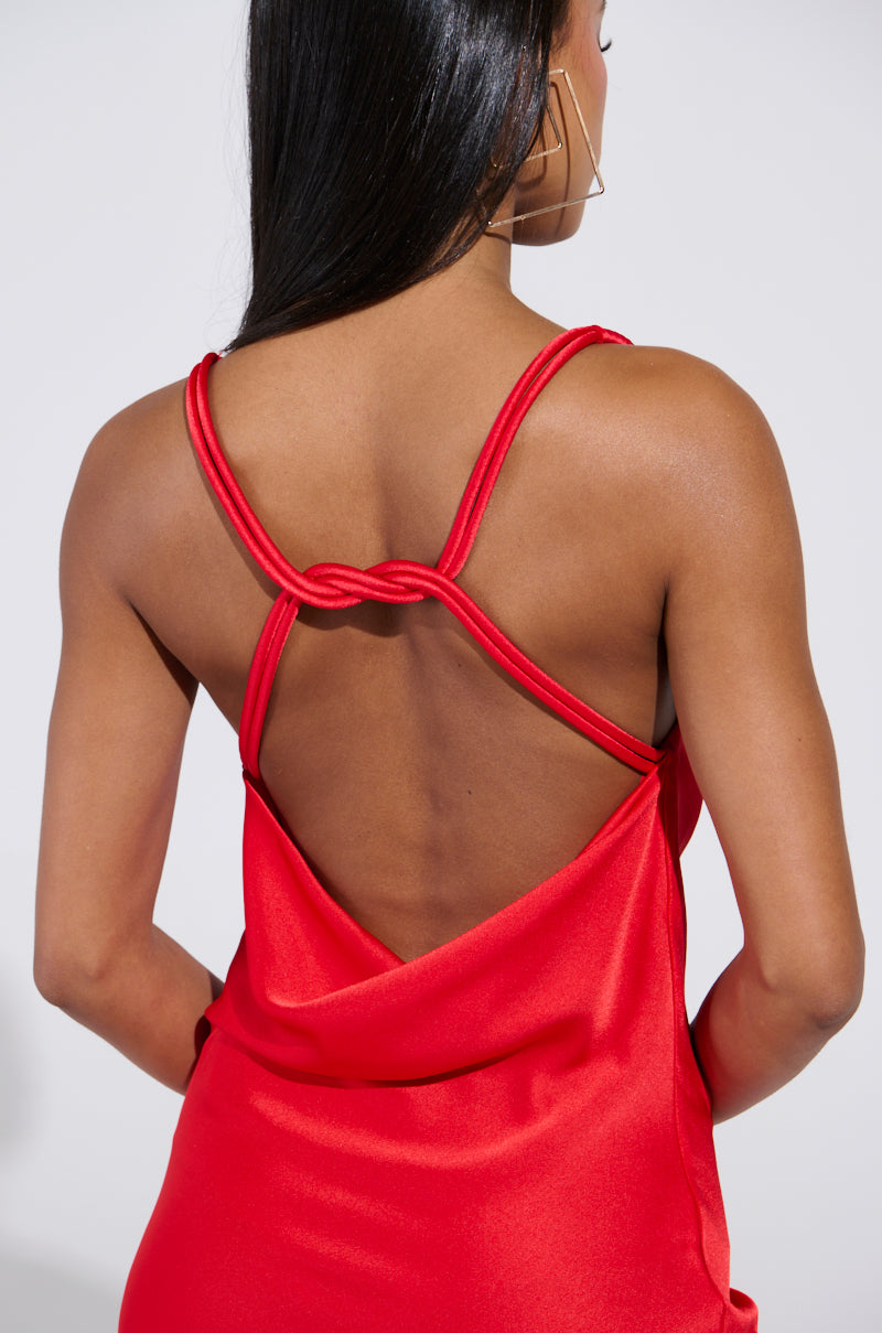 A woman with long dark hair is elegantly dressed in the BELLA SATIN MAXI DRESS IN RED, featuring a low open back with intricate looped strap details. She stands facing away from the camera against a light background.