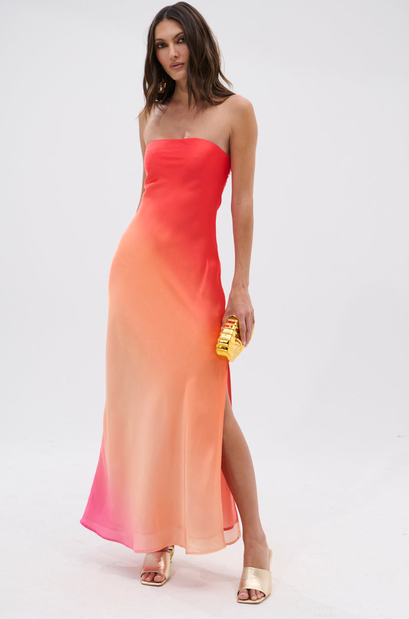A woman stands against a white background wearing the OCEAN BREEZE OMBRE SATIN MAXI DRESS, which fades from red to peach. She accessorizes with a yellow clutch and gold heeled sandals for an effortlessly chic, vacation-ready look.