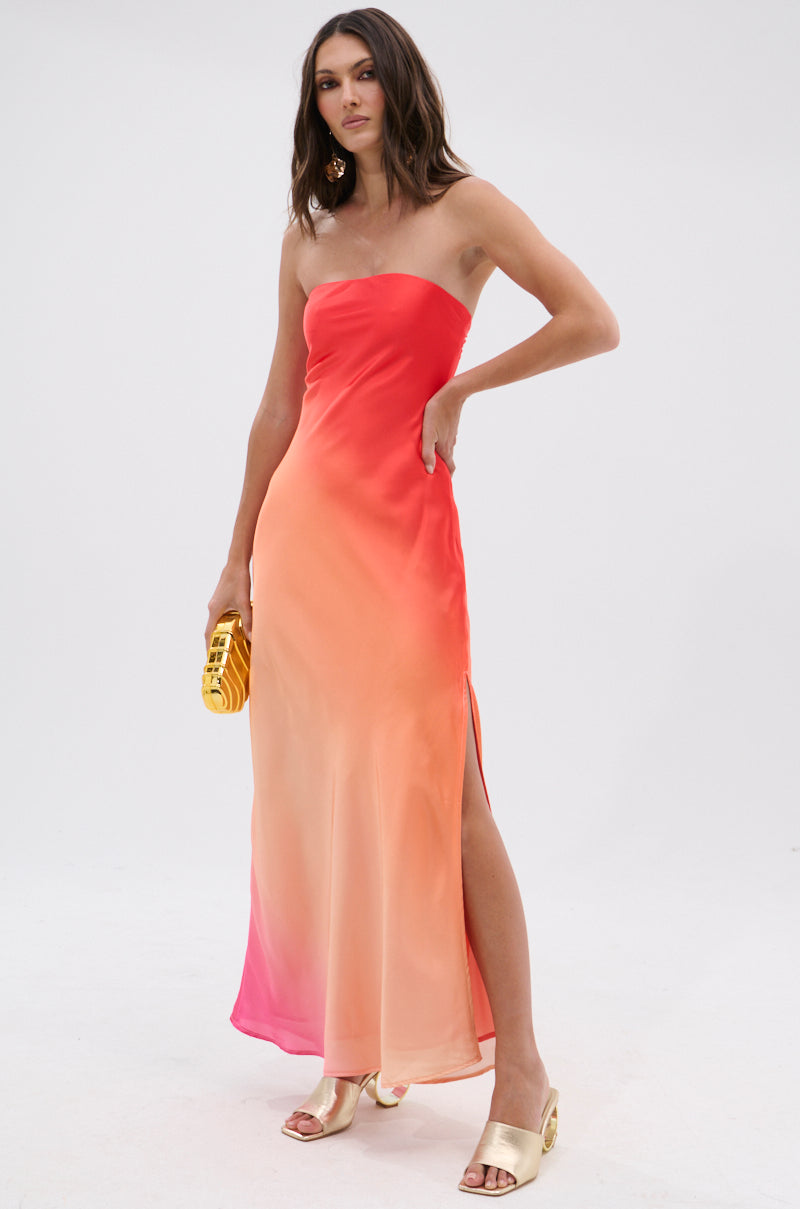 A woman in the OCEAN BREEZE OMBRE SATIN MAXI DRESS with a thigh-high slit, gold open-toe heels, and a small yellow clutch stands against a plain white background.