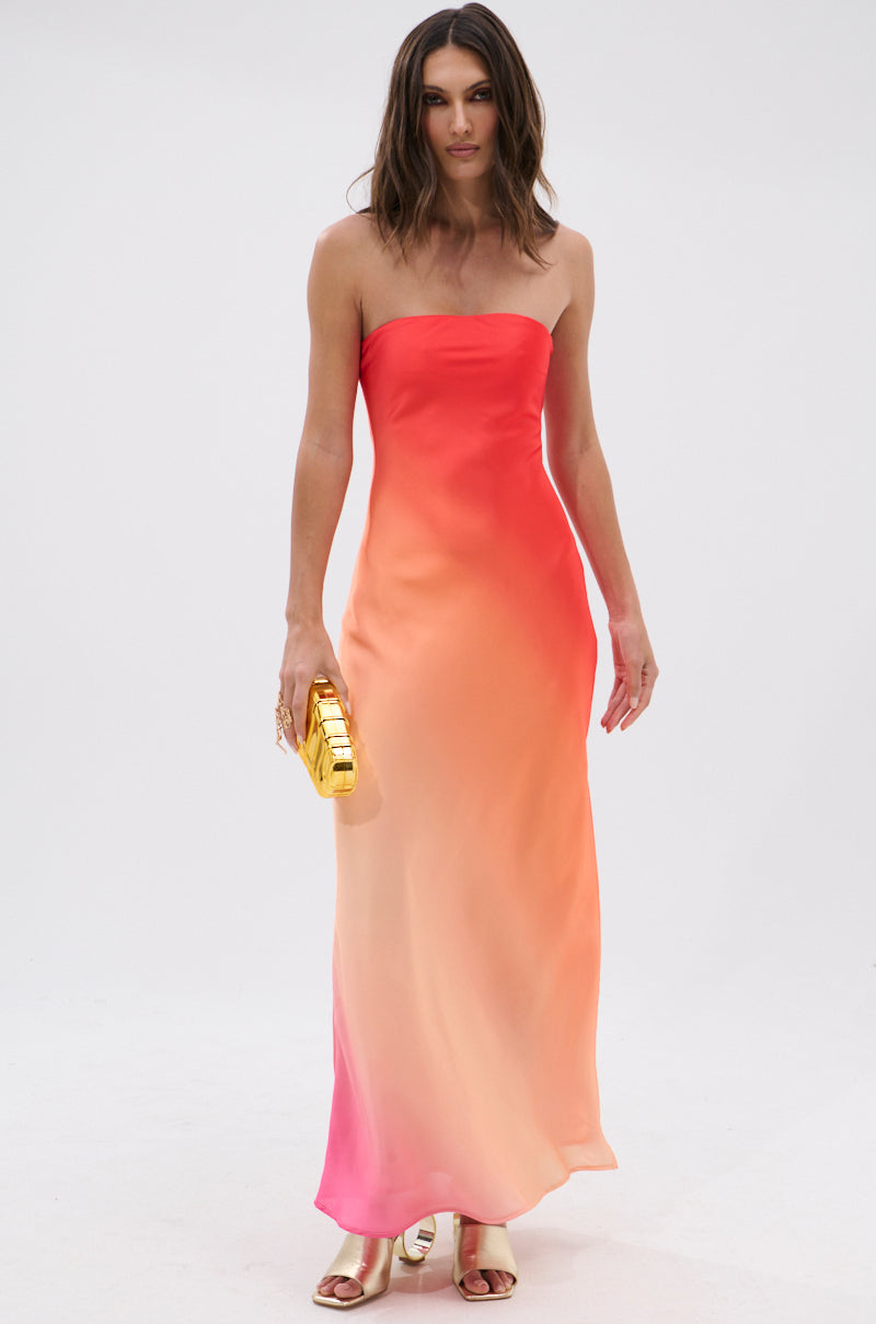 A woman wears the OCEAN BREEZE OMBRE SATIN MAXI DRESS, featuring a strapless design with coral-to-peach ombre satin. She pairs it with a gold clutch and gold open-toe heels, posing against a white background for a vacation-ready look.