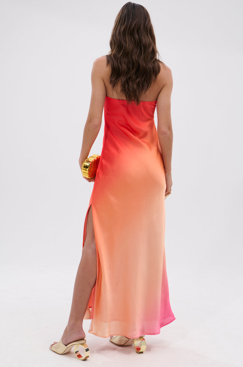A woman with wavy brown hair, seen from behind, wears the OCEAN BREEZE OMBRE SATIN MAXI DRESS in red, orange, and pink. She accessorizes with a small yellow handbag and cream-colored slide sandals for a vacation-ready look.
