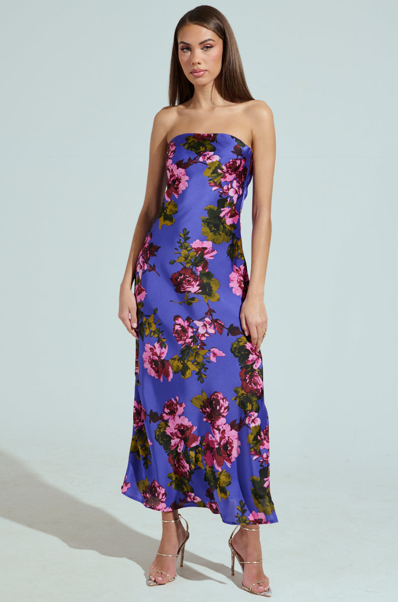A woman poses in the ROSE PETALS MAXI DRESS, a strapless blue satin gown adorned with vibrant pink and green florals. Her long straight hair cascades elegantly, complemented by chic high-heeled sandals against a plain backdrop.