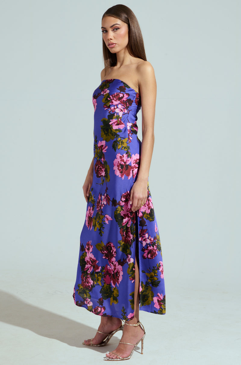 In a floor-length ROSE PETALS MAXI DRESS of purple satin with colorful floral prints, a person stands gracefully. Their long brown hair and sideways glance at the camera exude elegance against the plain light gray backdrop.