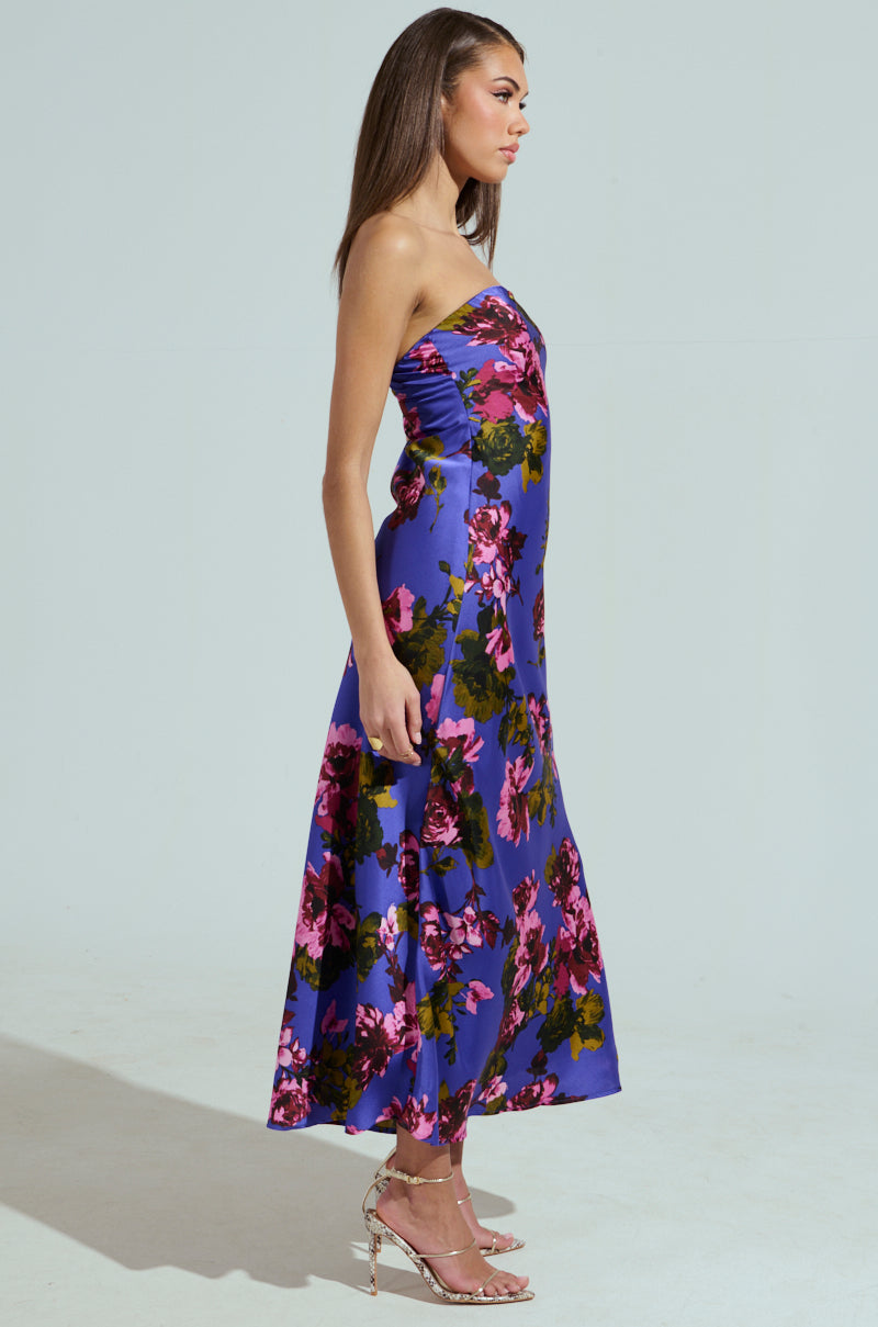 A woman in the ROSE PETALS MAXI DRESS, featuring a strapless floral satin design with vibrant pink and green patterns, stands in profile. Her long, straight hair and strappy high-heeled sandals complement the plain light gray backdrop, highlighting her elegant style.