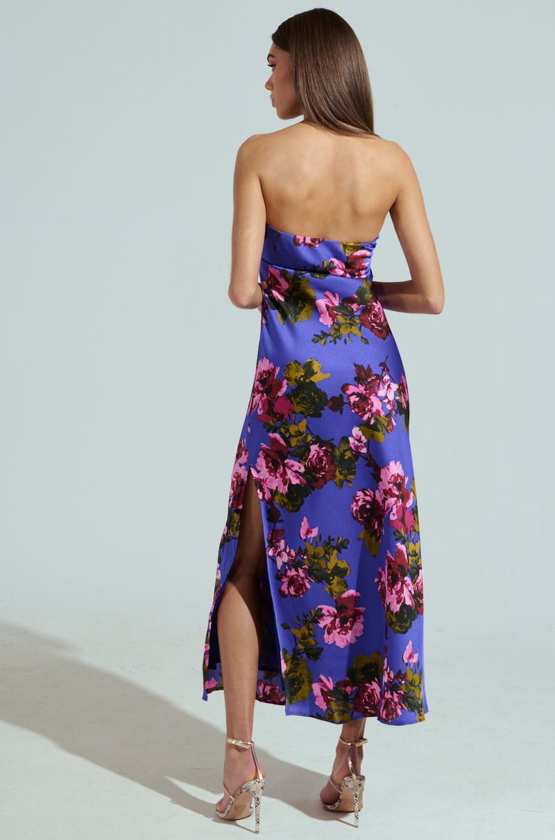 A woman stands with her back to the camera, wearing the ROSE PETALS MAXI DRESS, a strapless floral satin dress in blue with vibrant pink patterns. The maxi dress has a high slit and is elegantly paired with strappy high-heeled sandals against a plain light background.