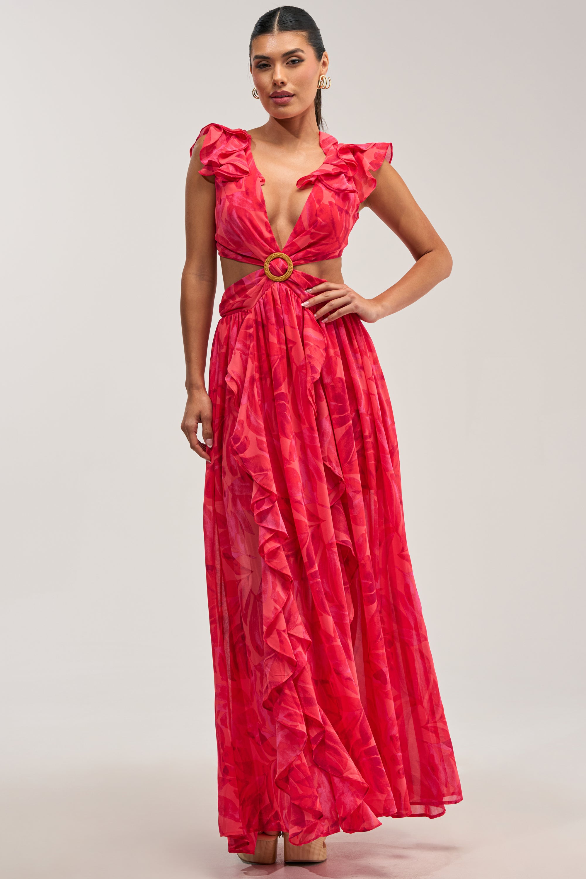 The FIRST CLASS FLORAL RUFFLE MAXI DRESS features a vibrant red floral print, deep V-neckline, chic ruffle trim, striking side cutouts with a gold ring buckle detail, and a flowing skirt—styled here with gold hoop earrings for an effortlessly chic summer date night ensemble.
