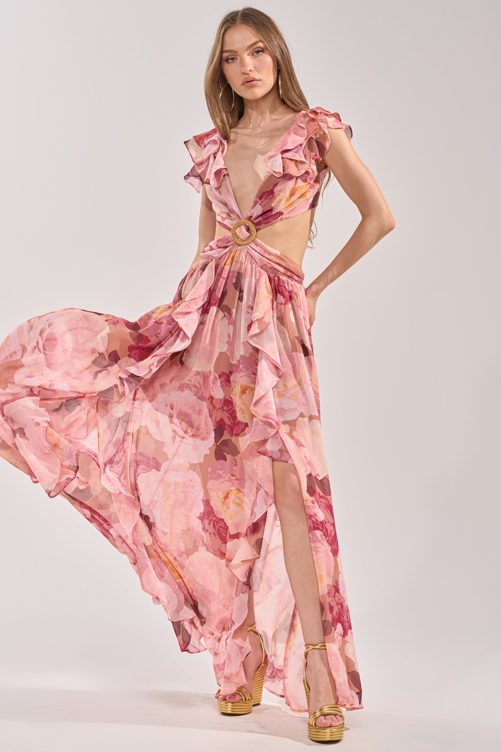 A woman models the LOVE YOU LATER FLORAL MAXI DRESS, featuring ruffles, a deep V-neck, cut-out sides, and a high slit. Styled with tan platform heels, she stands against a plain light background—ideal for spring occasions. 0