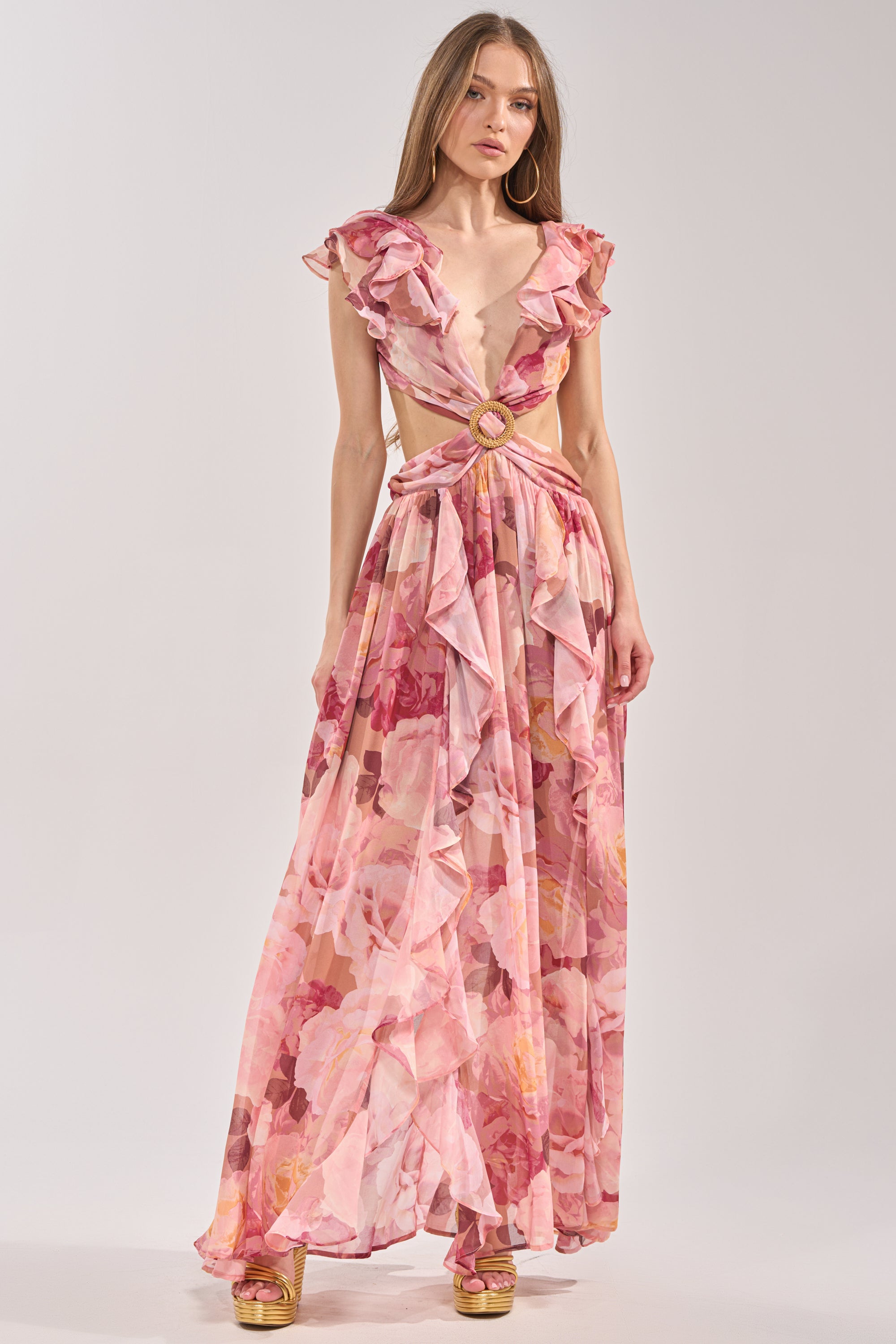 A woman wears the LOVE YOU LATER FLORAL MAXI DRESS, a pink maxi with ruffled sleeves and deep V-neckline, styled with gold platform sandals and long straight hair—ideal for spring occasions. 1