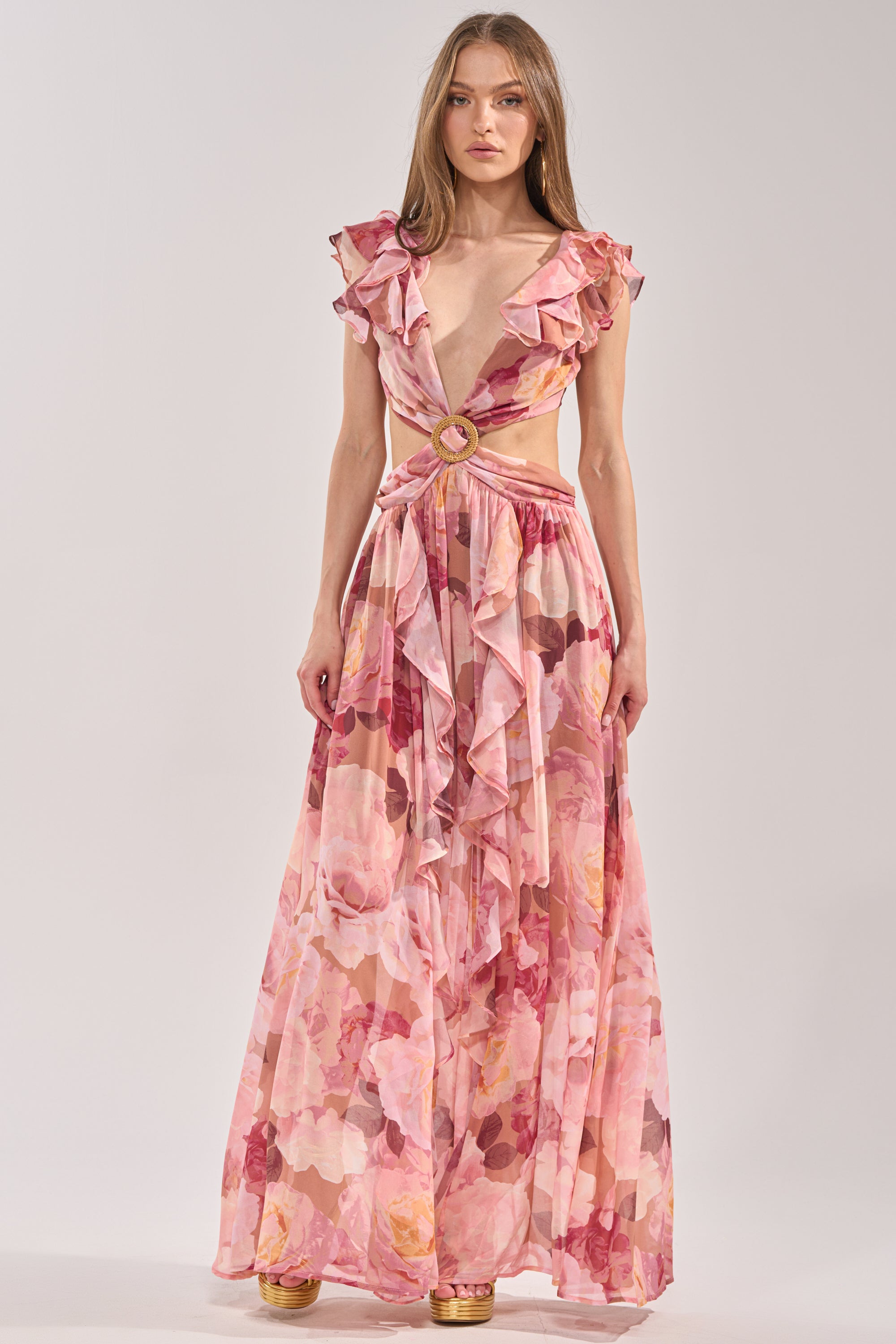 A woman stands against a plain background wearing the LOVE YOU LATER FLORAL MAXI DRESS, featuring a sleeveless, floor-length silhouette with ruffled shoulders, a deep V-neckline, gold ring at the waist, and flowy layered fabric. 2